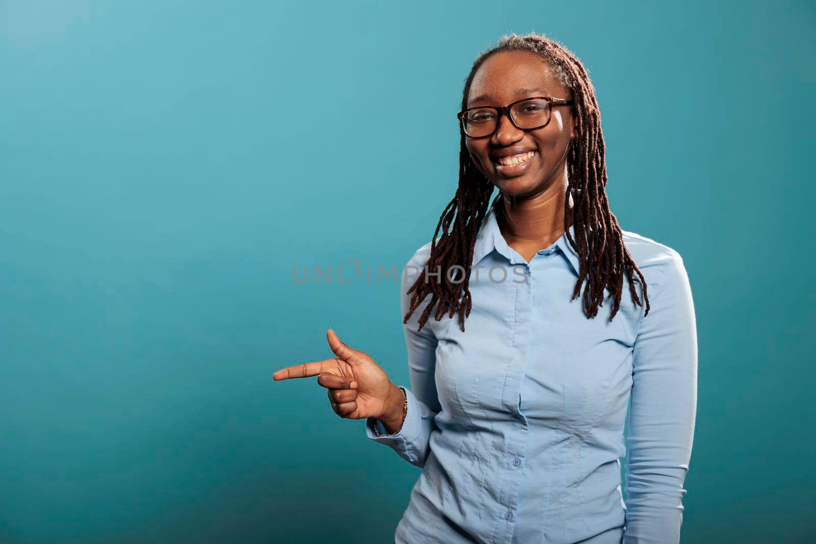 Happy excited young woman smiling heartily while pointing finger to left on blue background. Confident and positive adult person pointing hand to left while looking at camera.