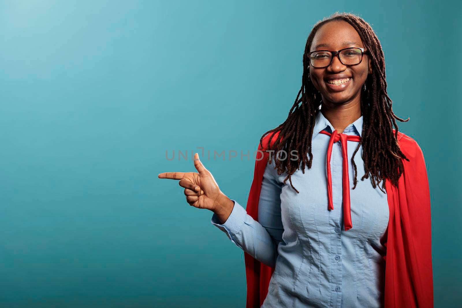 Joyful and confident superhero woman wearing red cape while pointing fingers to left on blue background. by DCStudio