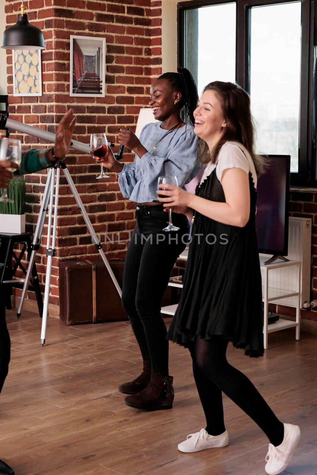 Smiling diverse women enjoying wine party event while dancing on funky disco music at home. Happy best friends celebrating friendship with crazy dance and alcoholic beverages.