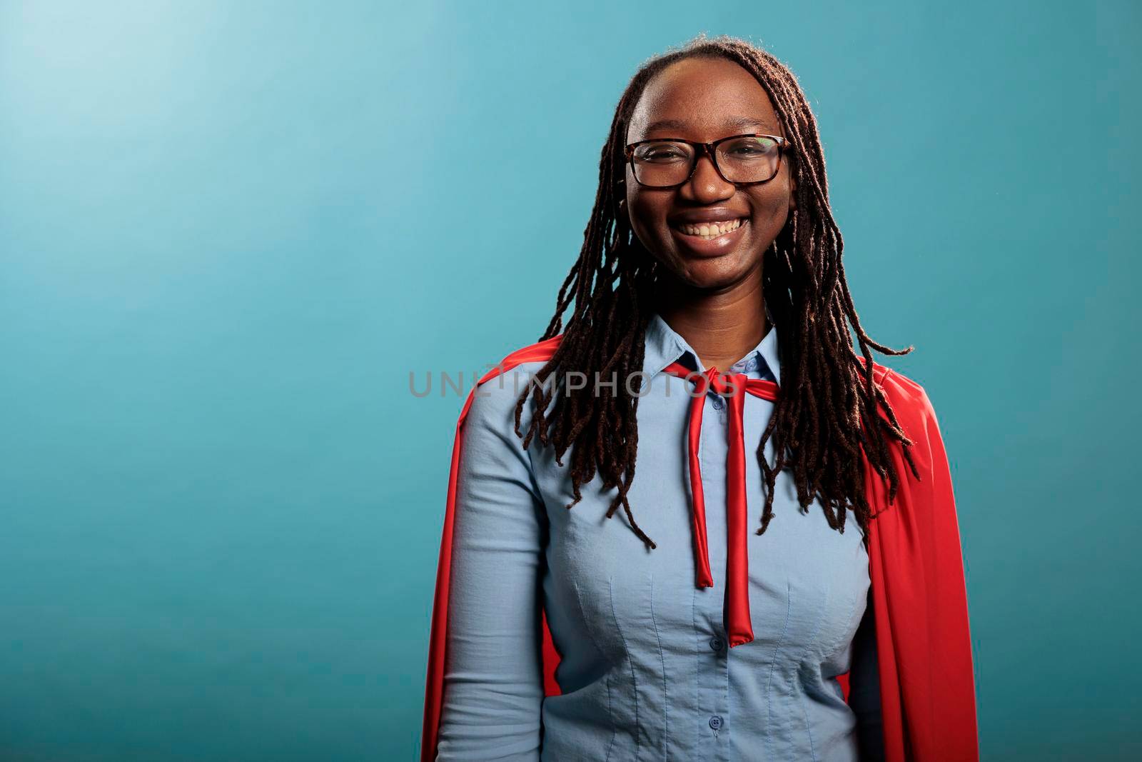 Portrait of african american young adult superhero woman wearing red cape on blue background while smiling at camera. by DCStudio