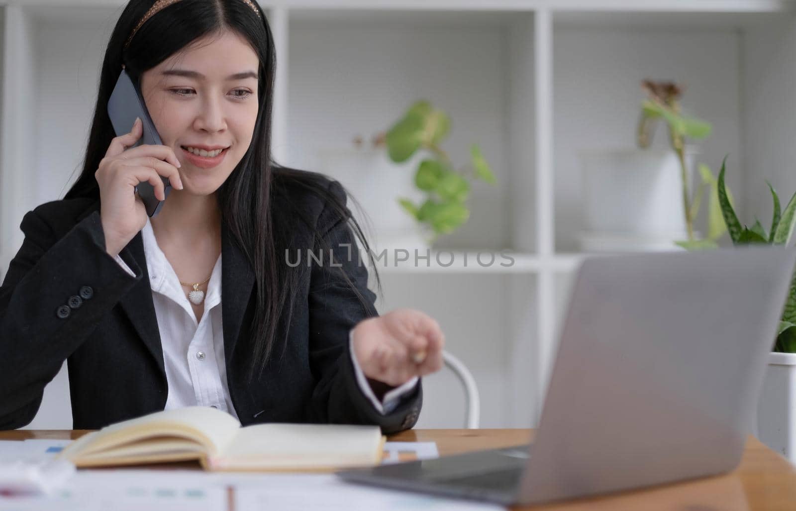 Smiling young Asian businesswoman sitting talking on the phone with laptop computer on desk in the office. by wichayada