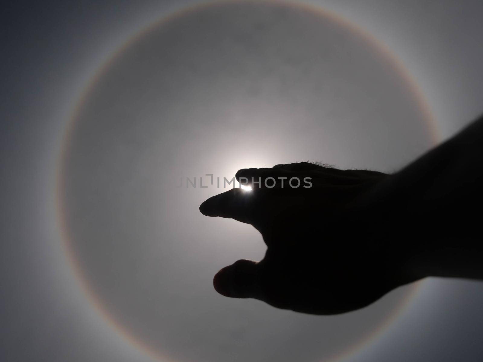 Beautiful photograph of the sun with a circular rainbow surrounded by a bright sky and white clouds with shadows of hands reaching out. Phenomenon, sun halo.