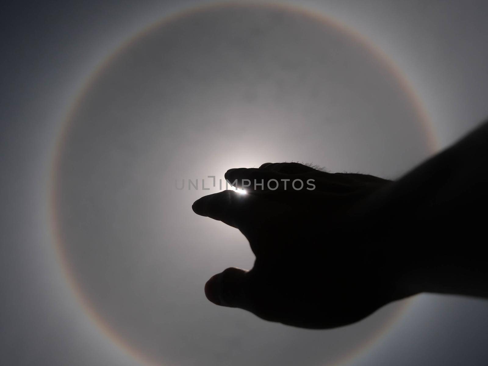 Beautiful photograph of the sun with a circular rainbow surrounded by a bright sky and white clouds with shadows of hands reaching out. Phenomenon, sun halo.