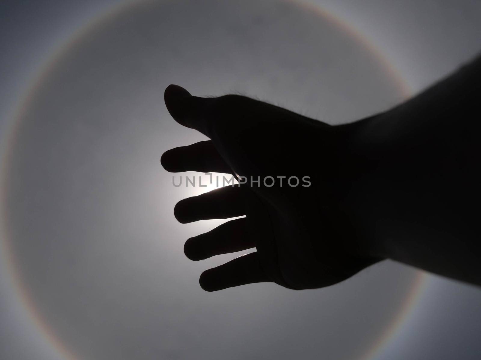 Beautiful photograph of the sun with a circular rainbow surrounded by a bright sky and white clouds with shadows of hands reaching out. Phenomenon, sun halo.