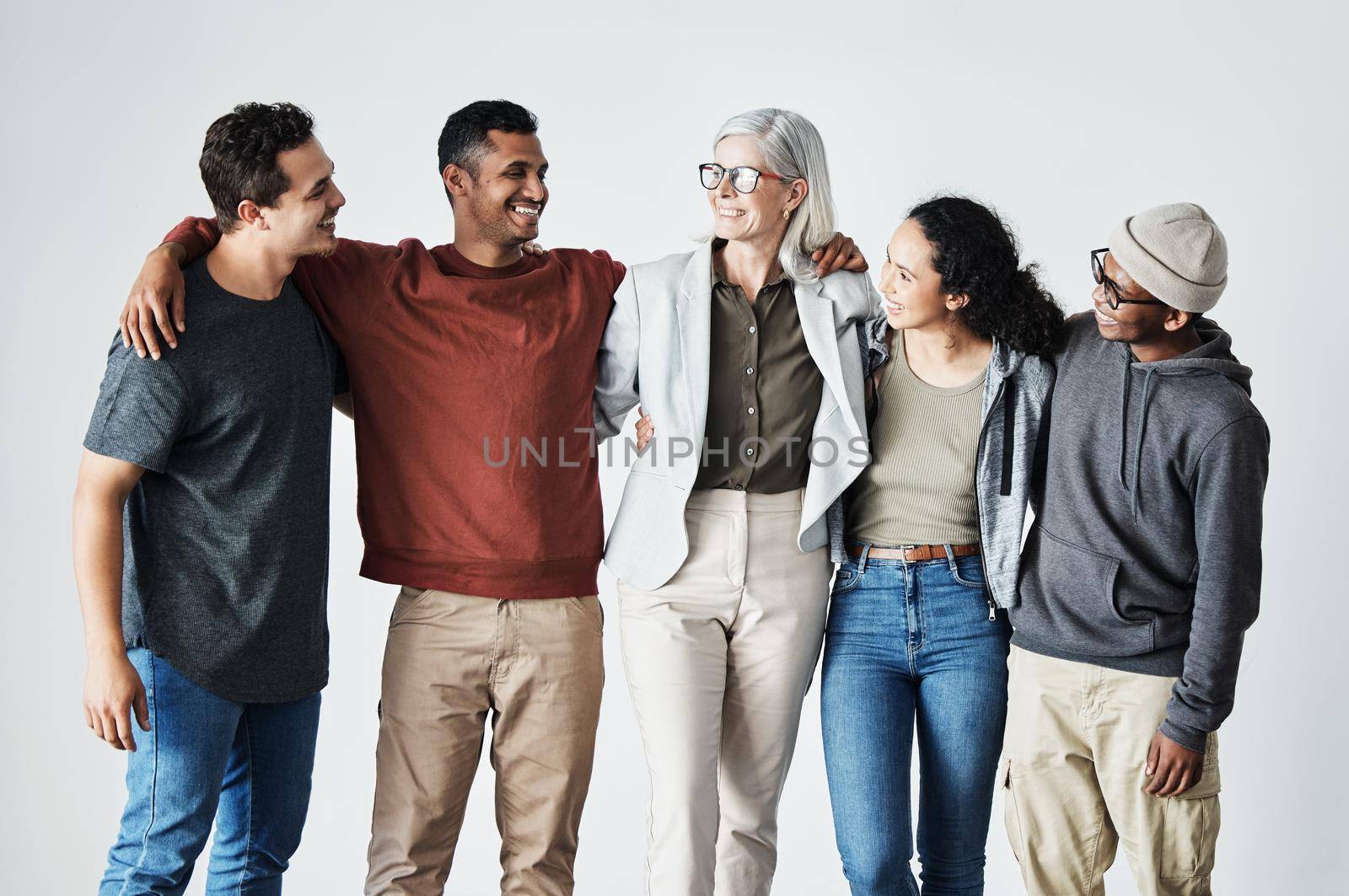 Team of happy united young business people hugging each other. Diverse mixed race group of men and women standing in row in their office, huddling and smiling all together. Unity and teamwork against a grey background by YuriArcurs