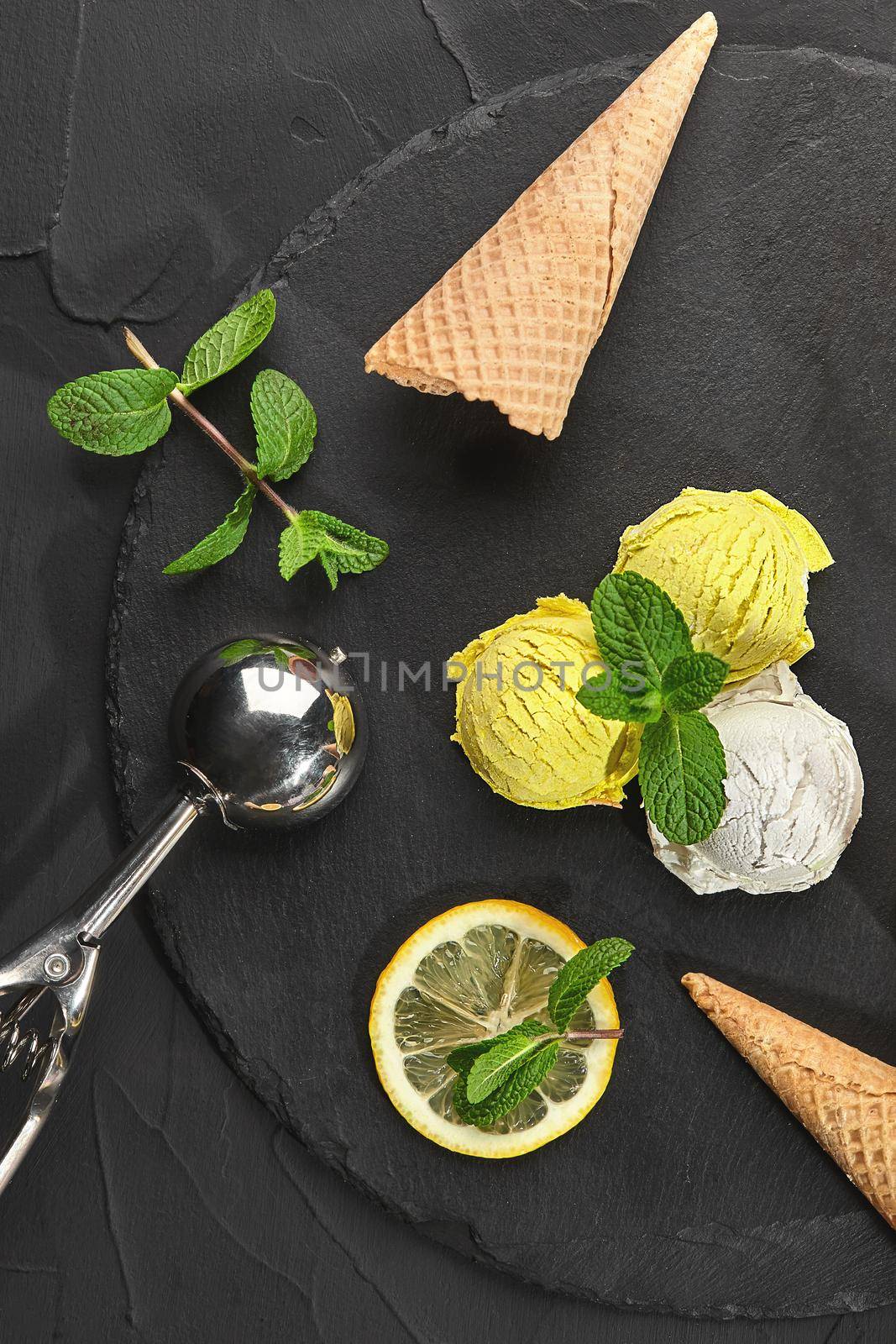 Close-up shot of a colorful creamy and lemon ice cream decorated with mint and served on a stone slate over a black background. Glossy scoop is laying nearby.