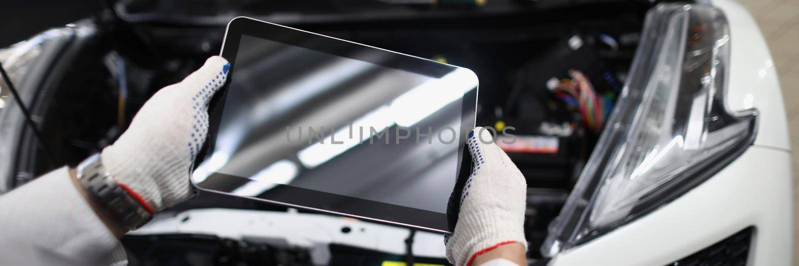 Close-up of mechanic use tablet device with black screen in front of car. Handyman using digital tablet at work in car service. Maintenance, mockup concept