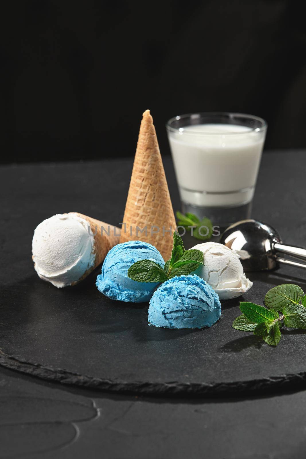 Close-up shot of a tasty natural creamy and blueberry ice cream decorated with fresh mint, and waffle cones are served on a stone slate, standing on a dark table over a black background. Metal scoop and a glass of milk is nearby.