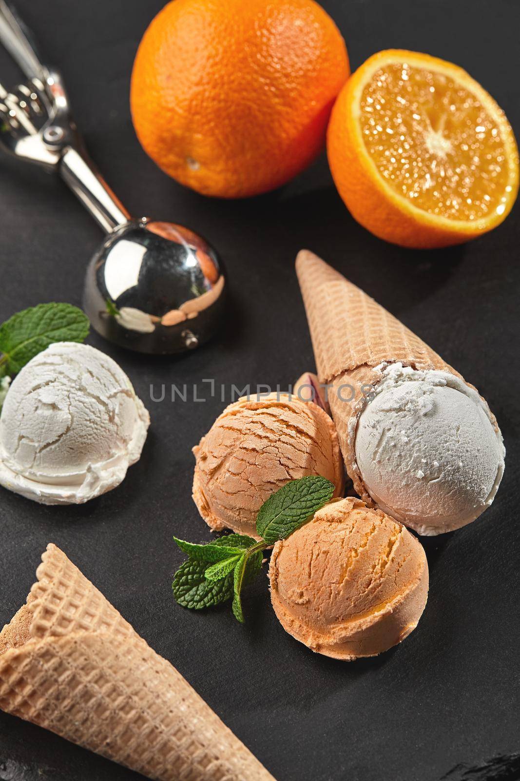 Stone slate tray with a delightful fresh creamy and orange ice cream set decorated with mint, and classic waffle cones on a dark table over a black background. Metal scoop is laying nearby. Summer coolness of ice cream and sorbet. Close-up shot.