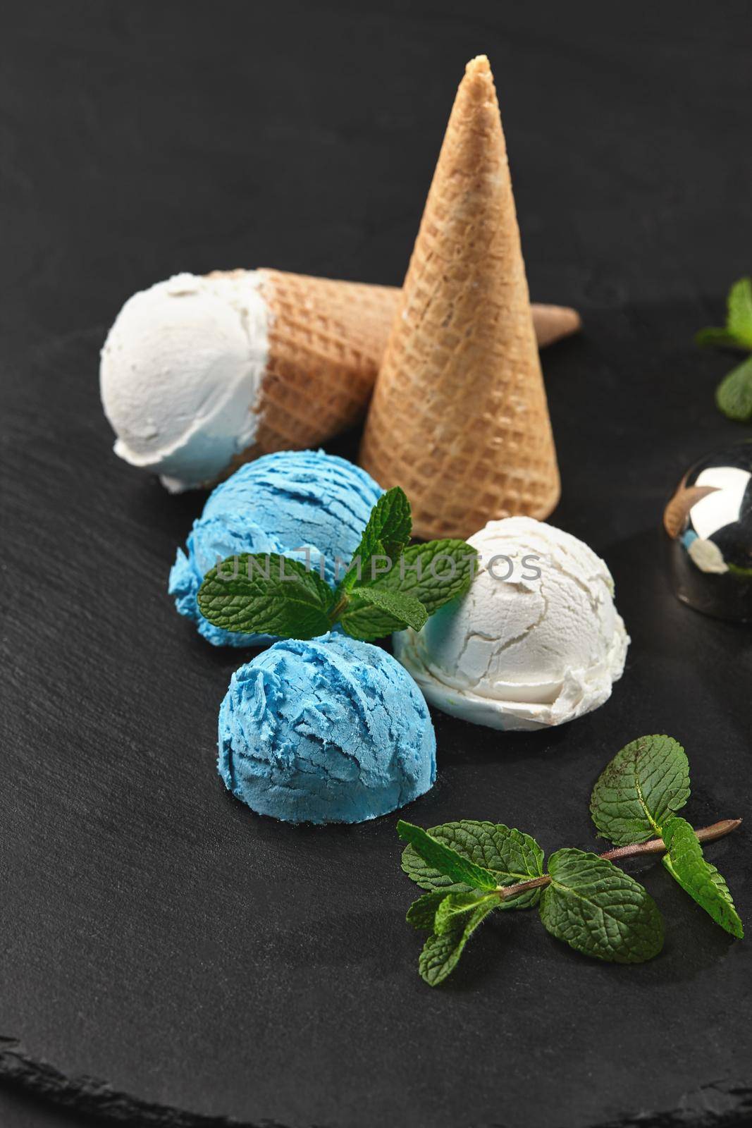 Top view of fantastic creamy and blueberry ice cream decorated with mint, and waffle cones are served on a stone slate standing on a dark table over a black background. Metal scoop is laying nearby. Close-up shot.