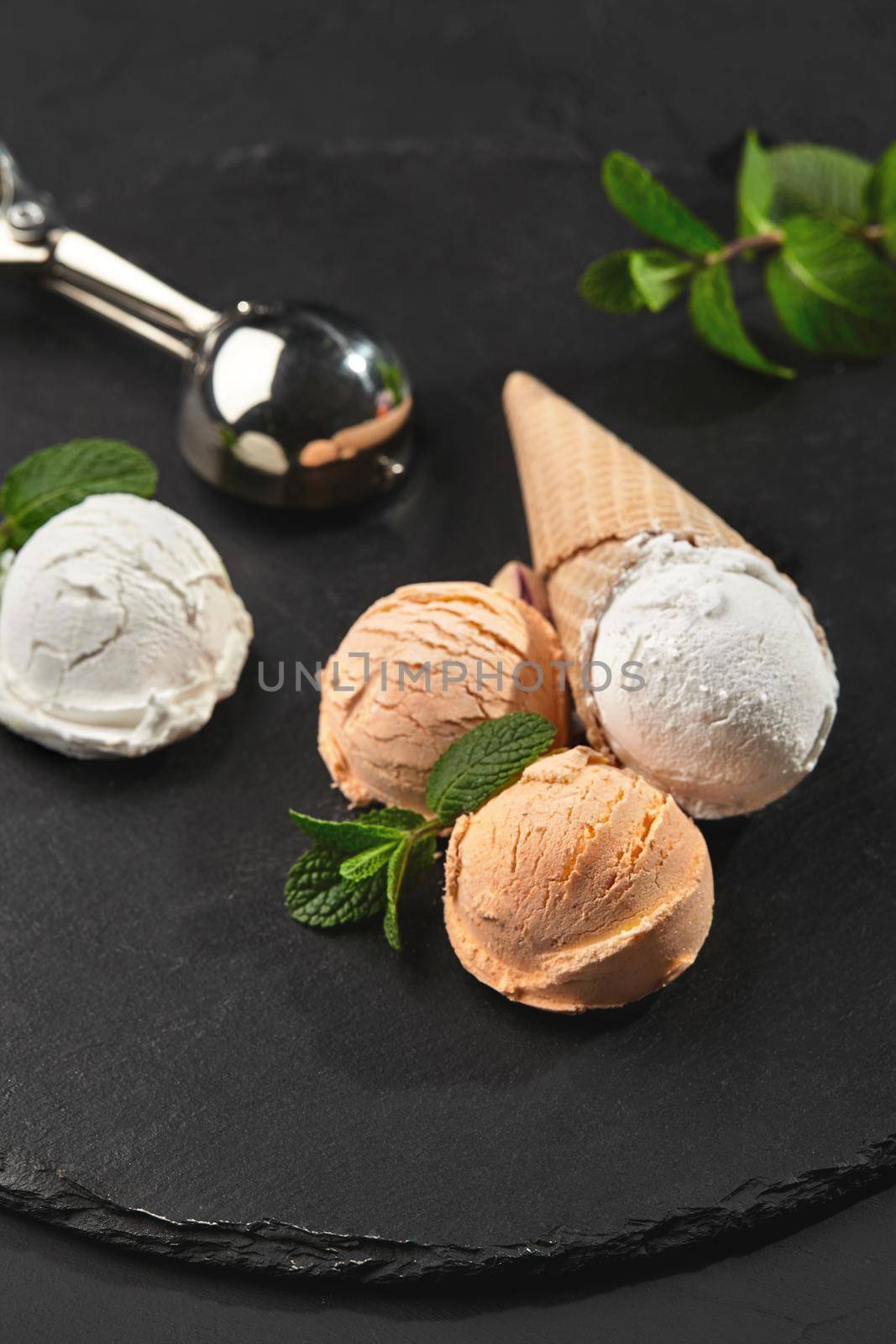 Stone slate tray with a tasty creamy and orange ice cream set decorated with fresh mint, and classic waffle cones on a dark table over a black background. Metal shiny scoop is laying nearby. Summer coolness of ice cream and sorbet. Close-up shot.