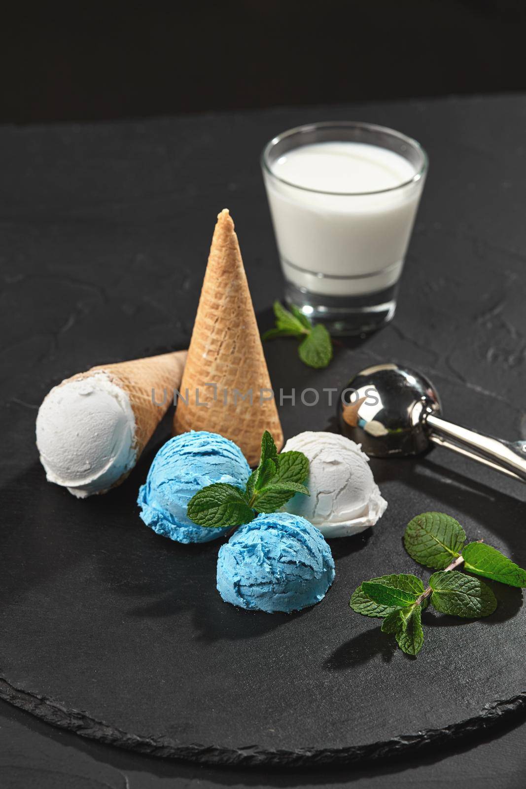 Top view of a delicious natural creamy and blueberry ice cream decorated with mint, and classic waffle cones are served on a stone slate, standing on a dark table over a black background. Metal scoop and a glass of milk is nearby. Close-up shot.