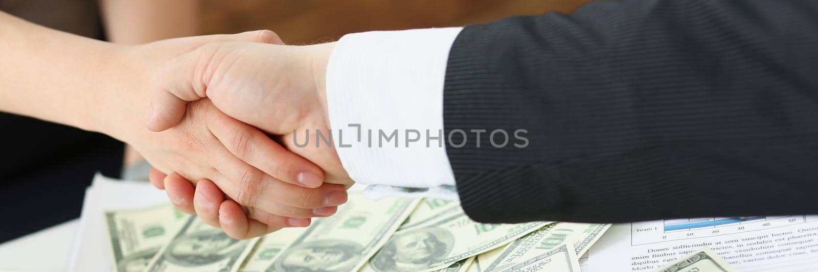 Close-up of businesspeople shake hands over bunch of dollar banknotes, transaction of powers. People agreed on price. Business, money, investment concept