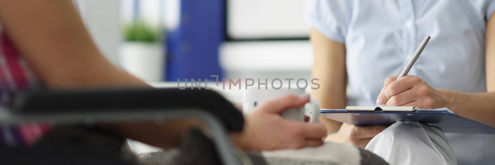 Close-up of doctor hold clipboard with patient medical history on appointment. Female ask questions disabled client in wheelchair. Nursing house concept