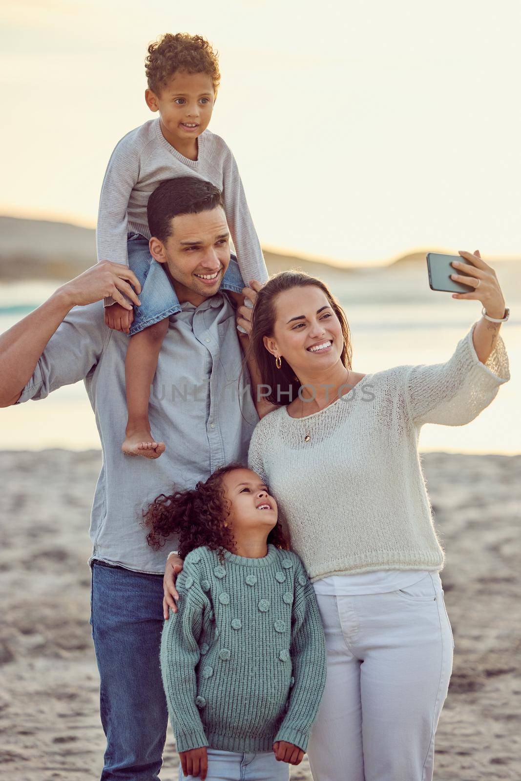 Happy cheerful mixed race family smiling for a selfie spending time at the beach together. Hispanic mother smiling taking a photo with her children and husband with her cellphone bonding on vacation by YuriArcurs