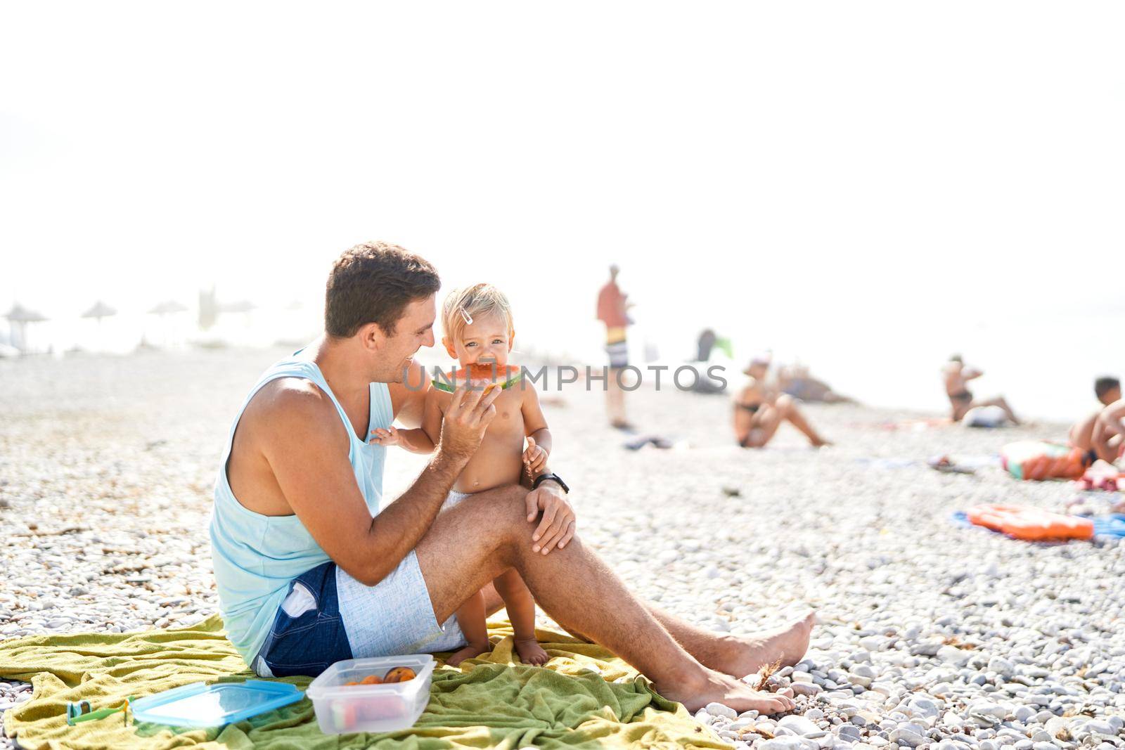 Smiling dad feeding little girl with slice of watermelon on beach. High quality photo