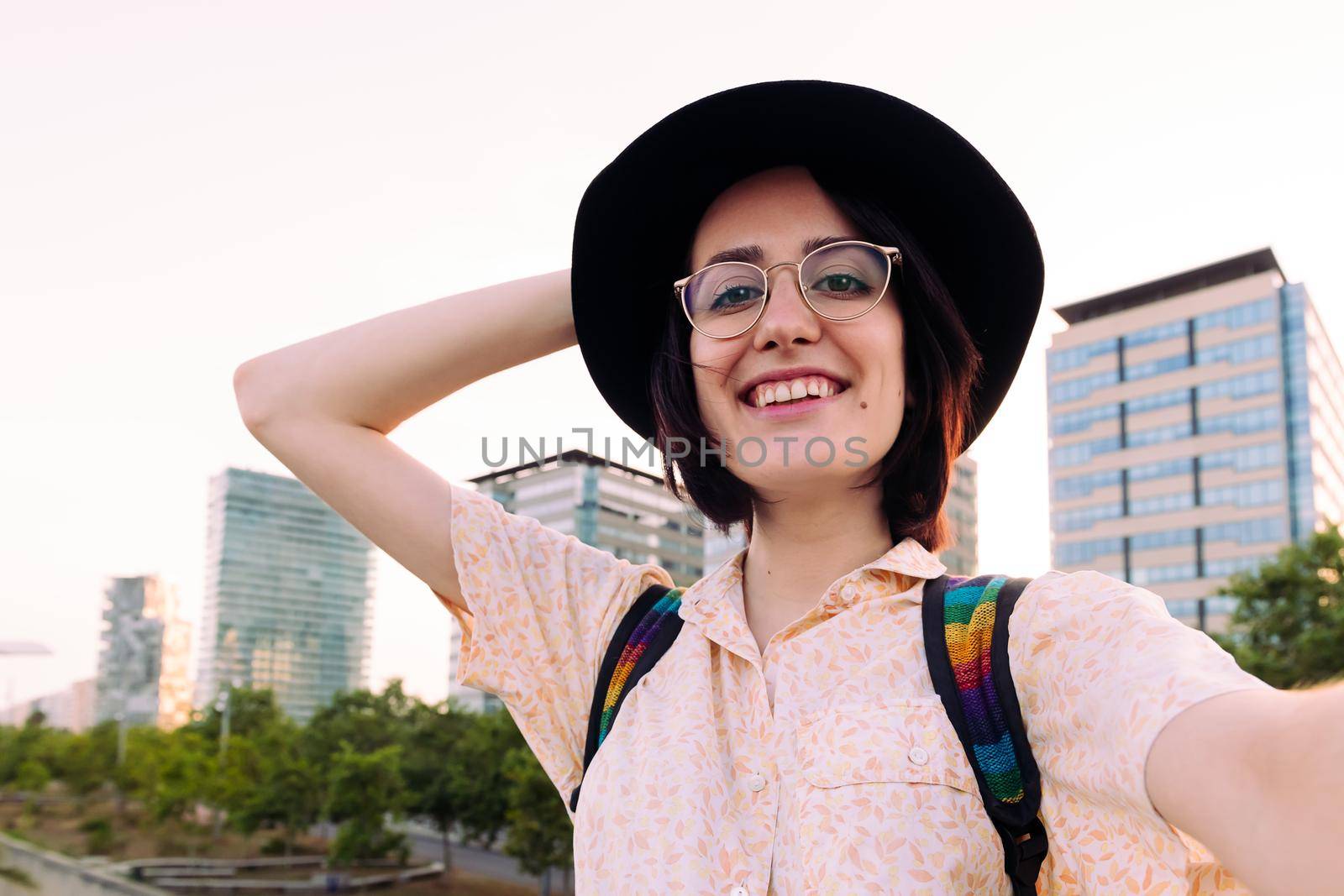 selfie photo of a beautiful young woman with glasses and hat smiling in front of the city, concept of social media and traveler lifestyle, copy space for text