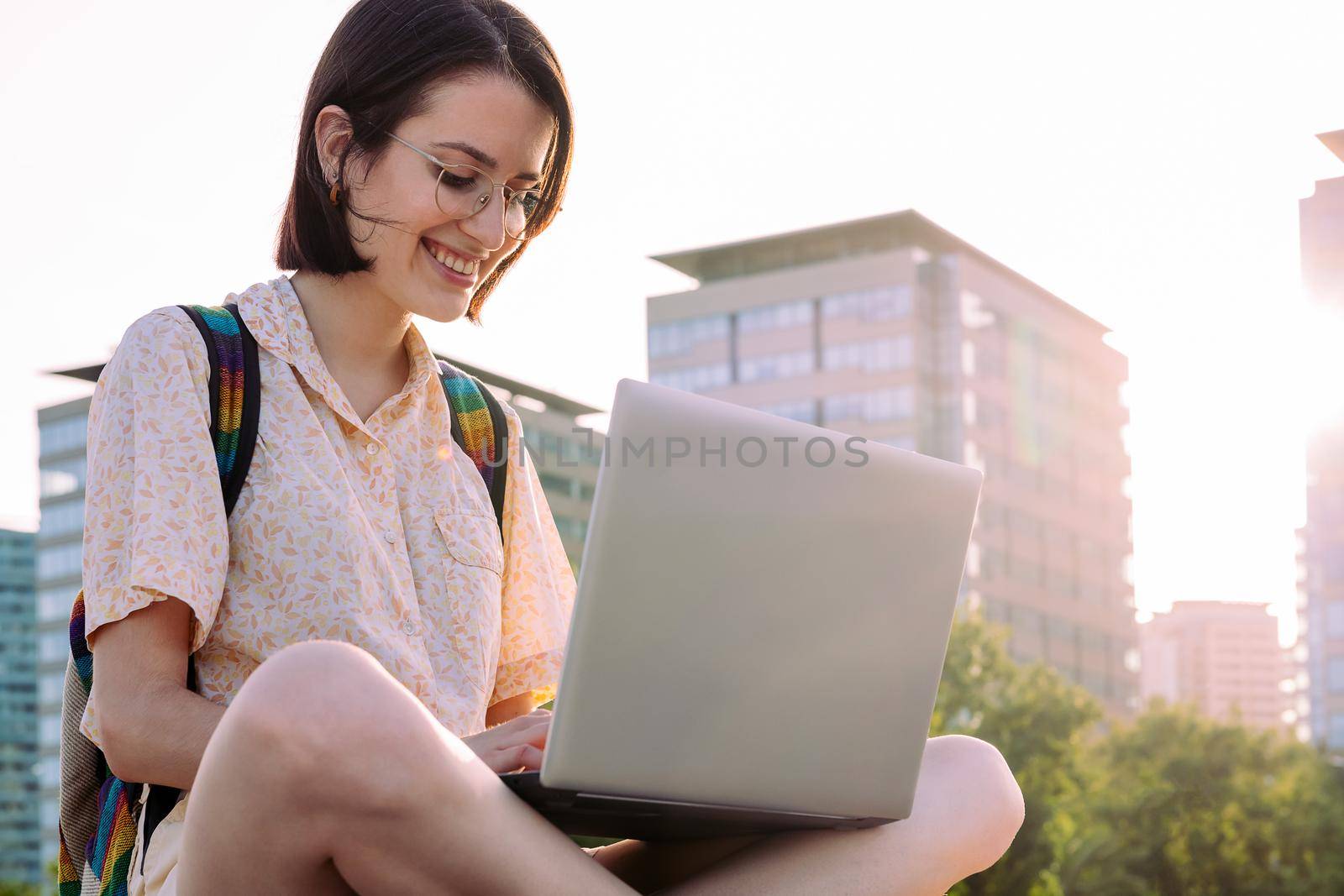 smiling young traveler woman with backpack sitting in the city typing on her laptop, concept of technology, youth and digital nomad lifestyle, copy space for text