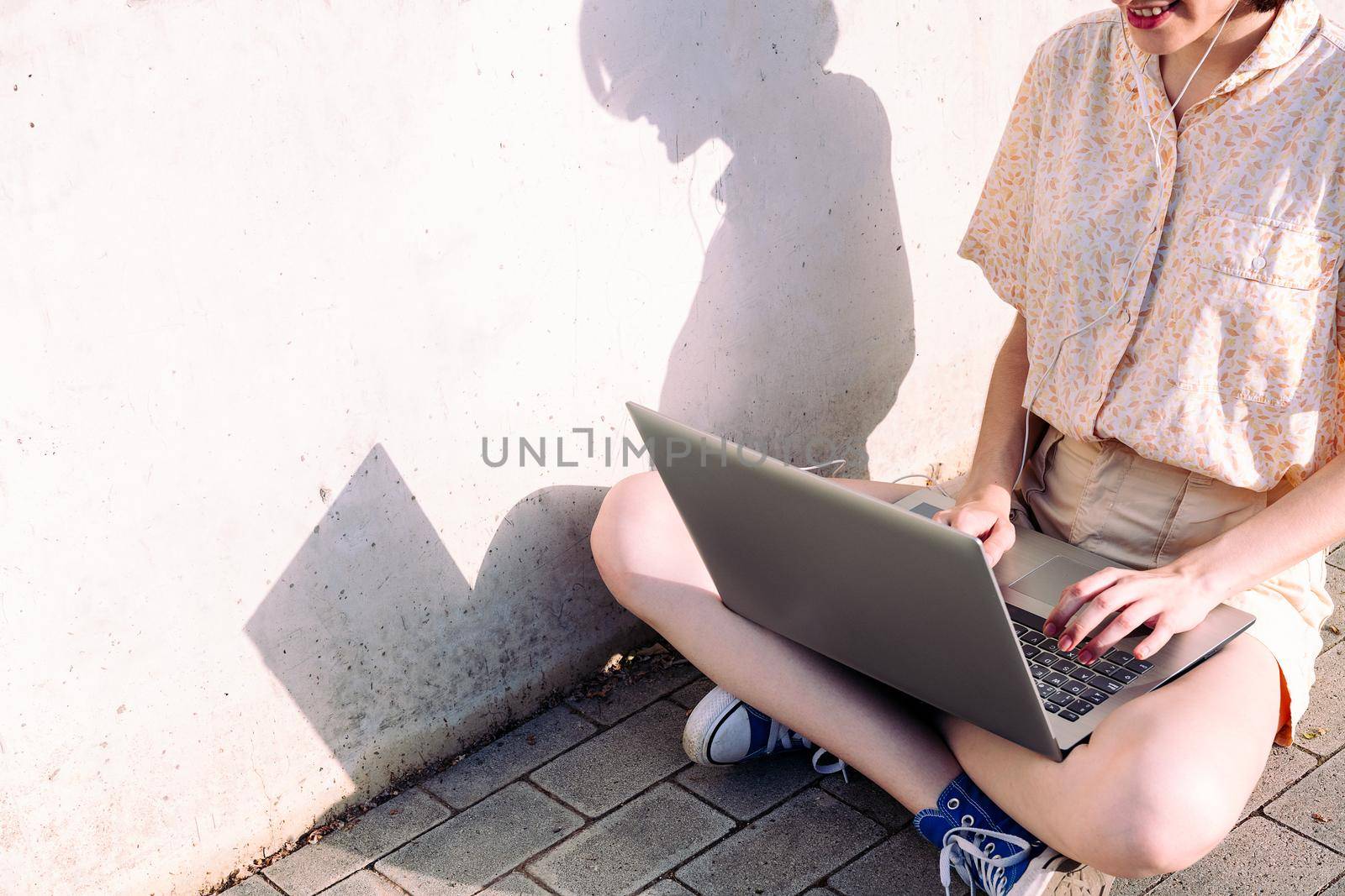 unrecognizable young woman with earphones working with a computer sitting next to a gray wall, technology concept and blogging lifestyle, text copy space