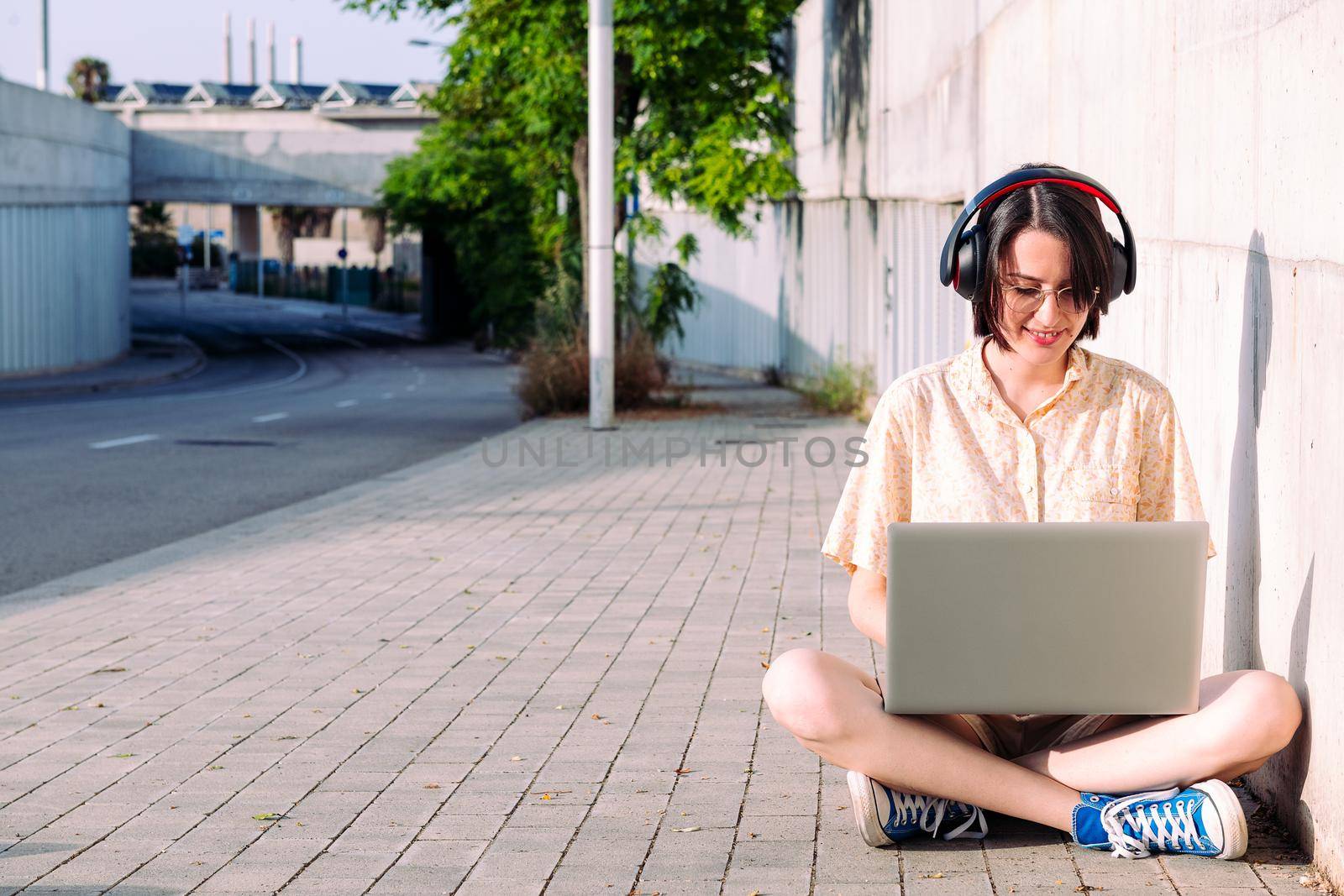 front view of a young woman with headphones working with a computer sitting next to a gray wall, technology concept and urban lifestyle, text copy space