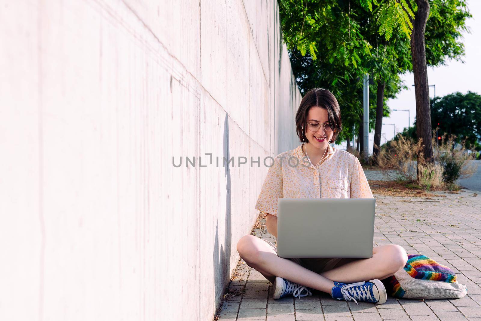 front view of a young woman with earphones working outdoors with a computer sitting next to a gray wall, technology concept and blogging lifestyle, text copy space