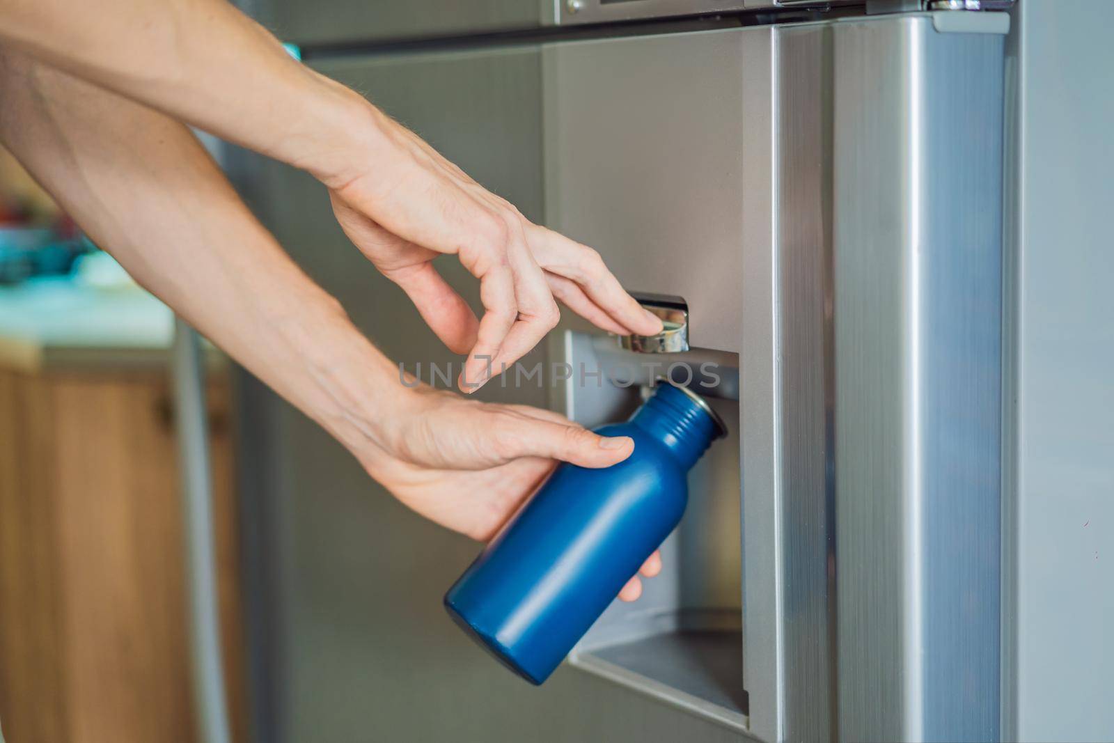 Male hand is pouring cold water and ice cubes in a metal bottle from dispenser of home fridge by galitskaya