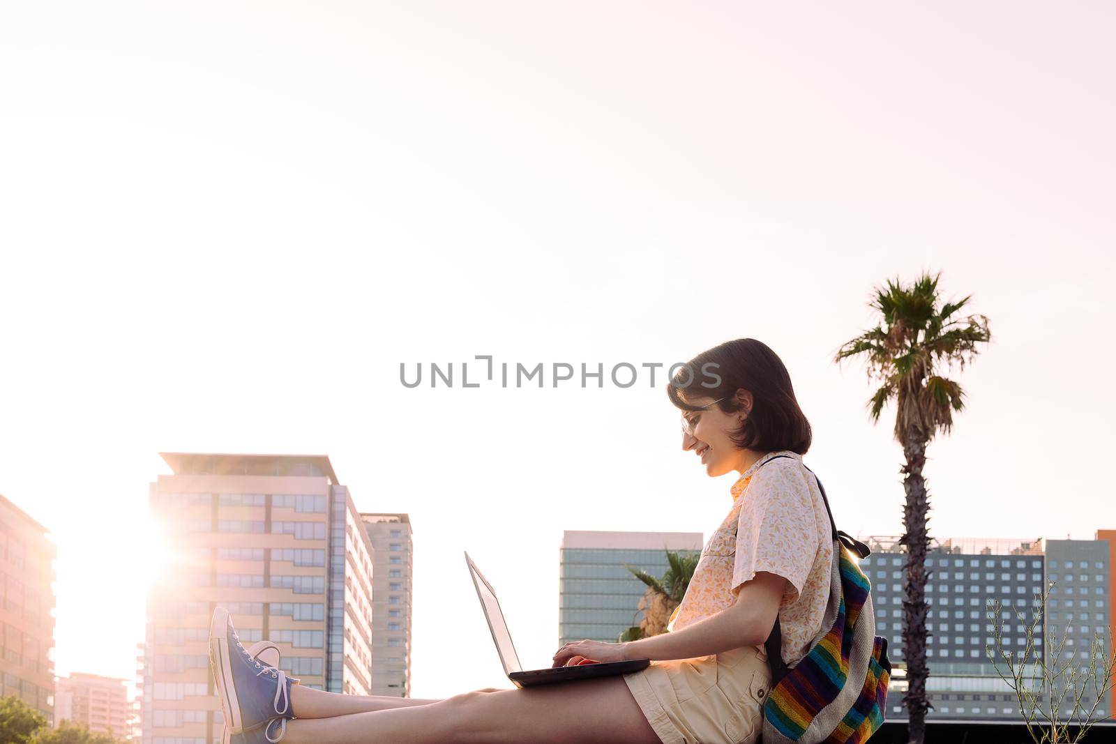 happy young traveler woman smiles working in the city at sunset with her laptop and backpack, concept of technology, youth and digital nomad lifestyle, copy space for text