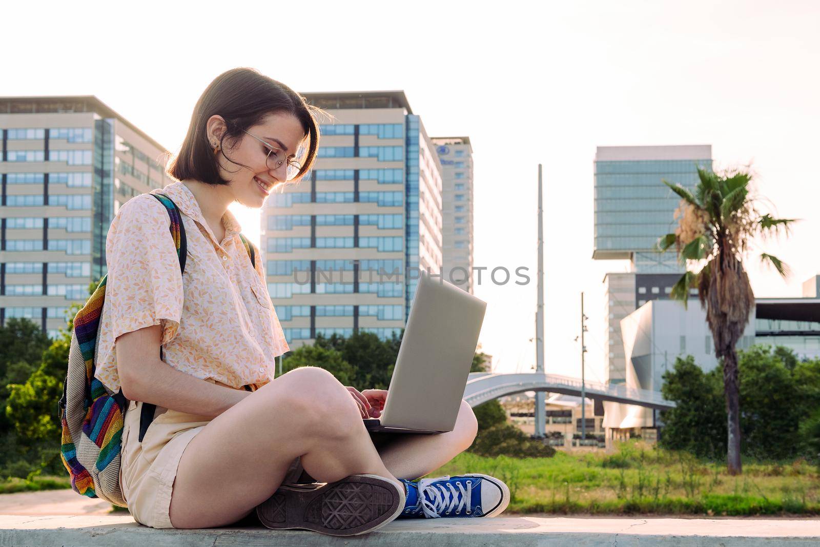 young traveler woman with backpack sitting in the city typing on her computer, concept of technology, youth and digital nomad lifestyle