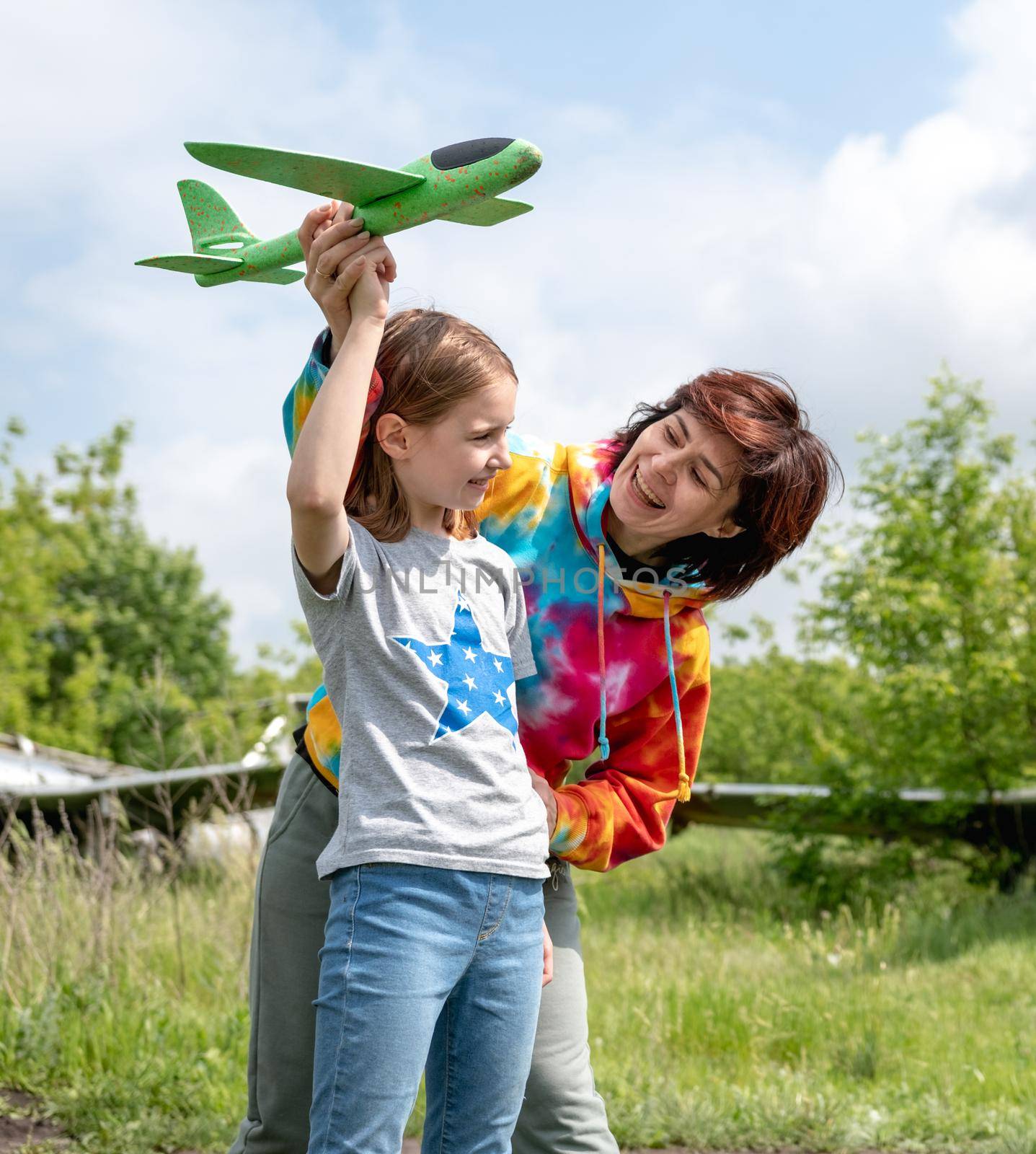 Mother and daughter child playing with toy plane at the field together with real airplane on background and smiling looking at each other. Happy family moments at the nature