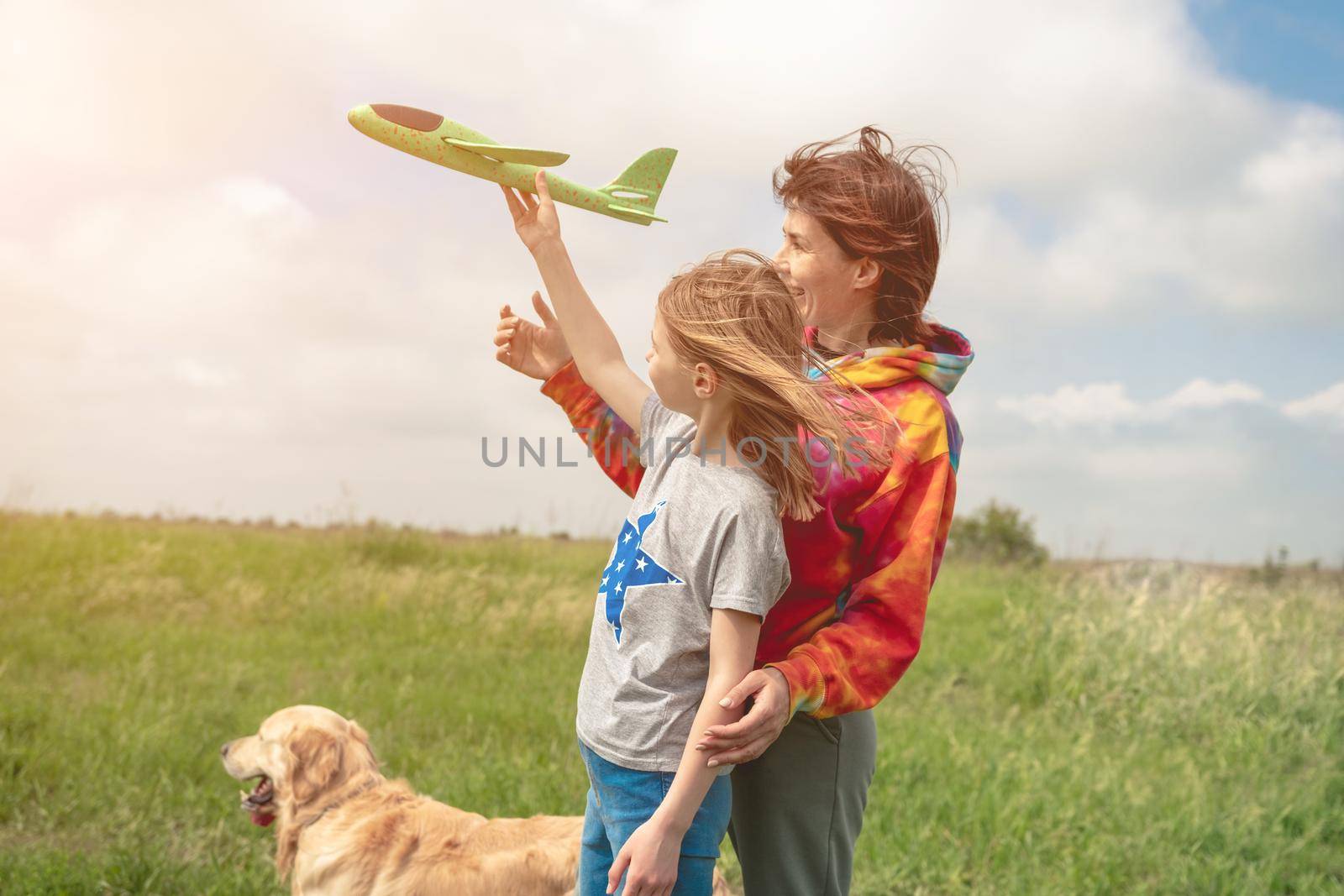 Mother and daughter launching toy plane to the sky and golden retriever dog close to them spending time at the field. Girl, woman and pet doggy playing with airplane at the nature