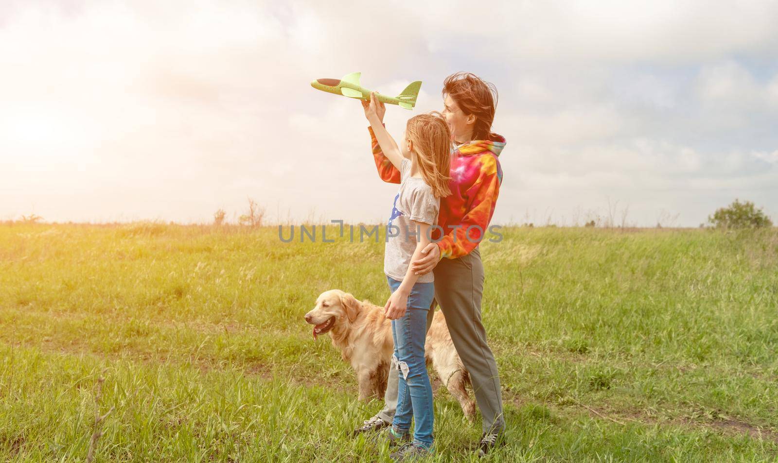 Mother and daughter with toy plane and golden retriever dog spending time at the field together. Girl, woman and pet doggy playing with airplane at the nature