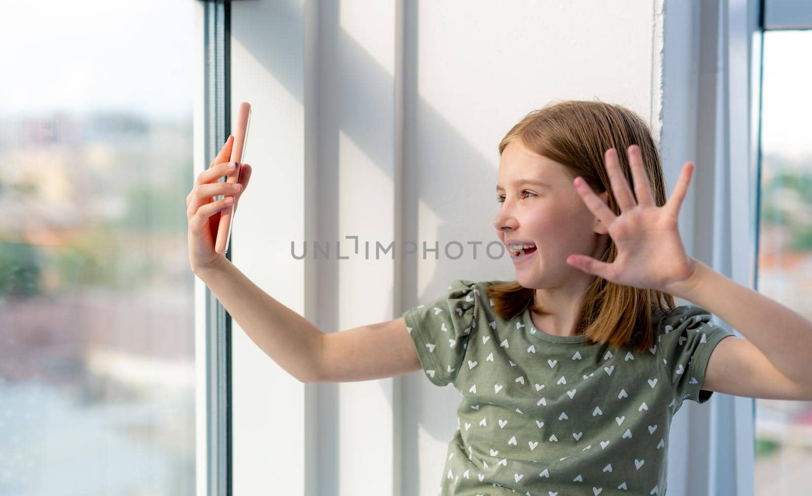 Adorable pretty preteen girl holding smartphone and making selfie with daylight from window. Beautiful female kid schoolgirl posing with cell phone at home