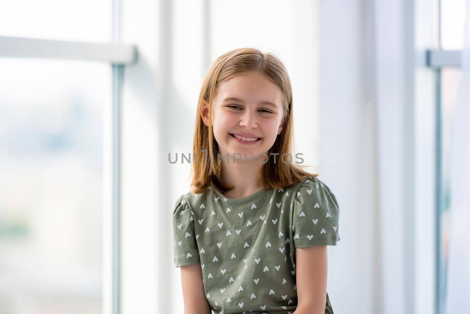 Portrait of preteen beautiful blond hair girl in light room with windows. Female kid schoolgirl looking at the camera and smiling indoors