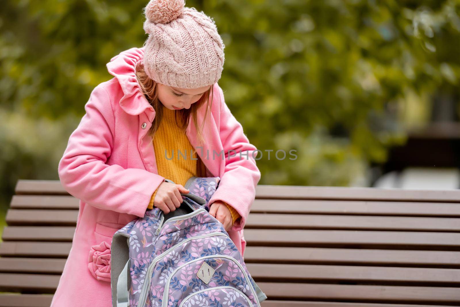 School girl kid looking at backpack sitting on the banch at autumn park. Beautiful female child resting after lessons