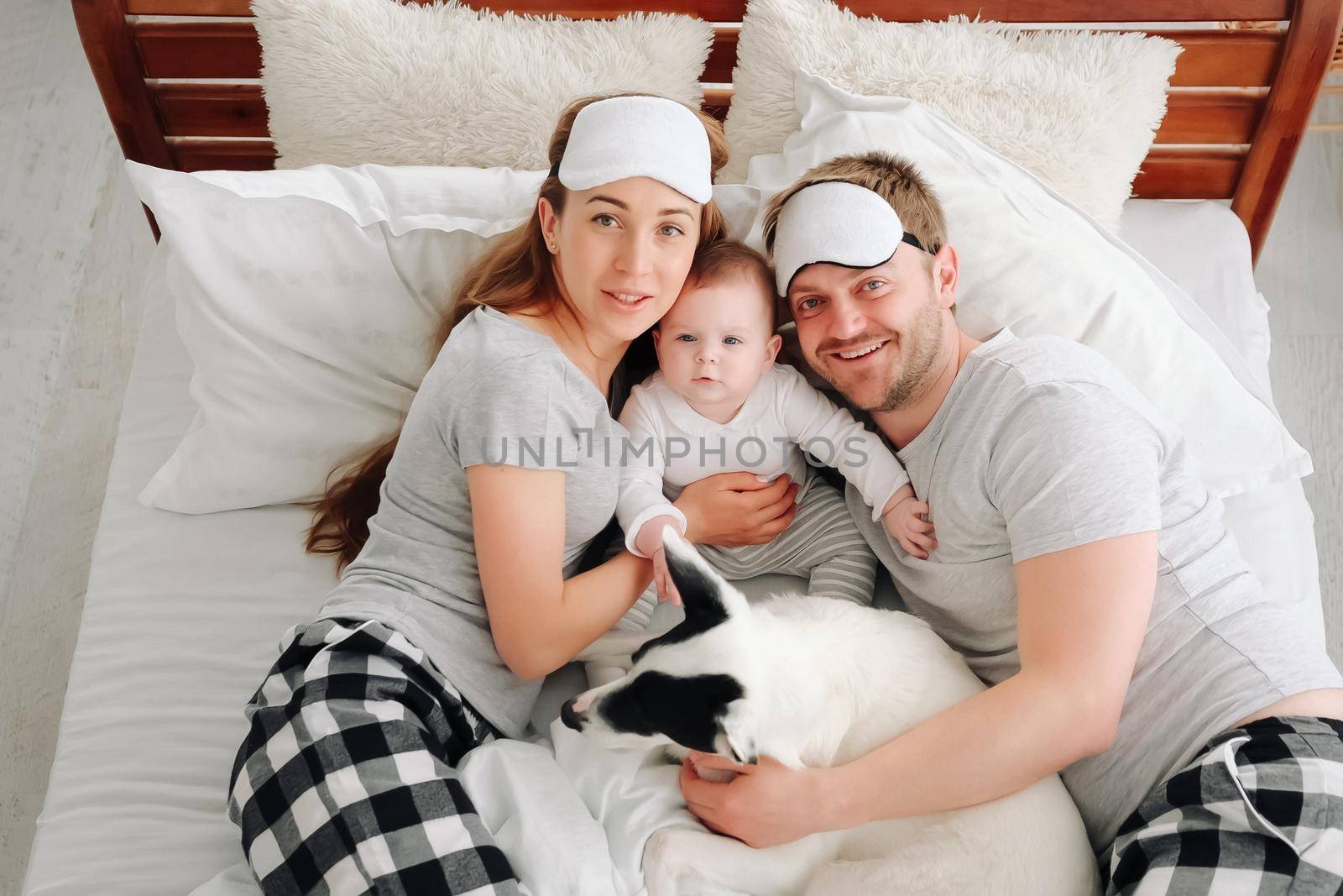Family with child and dog lying in the bed together, looking at the camera and smiling. Young mother and father hugging toddler kid and pet doggy in the bedroom