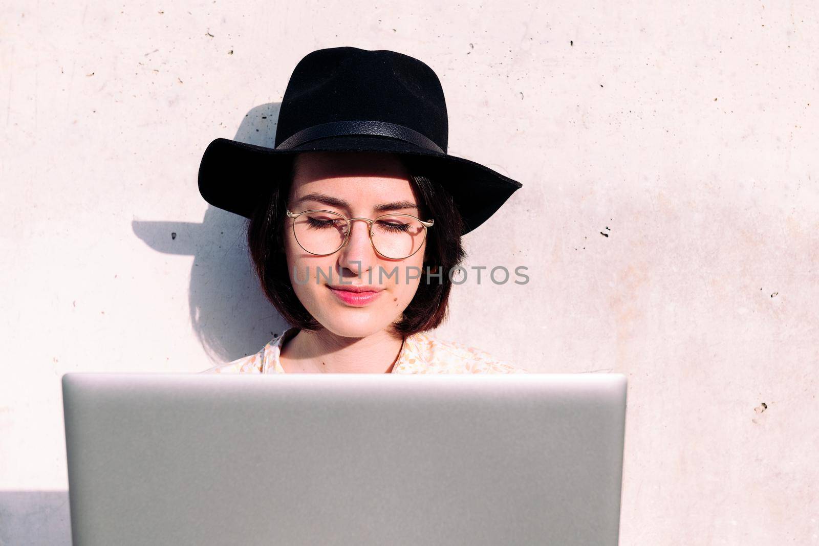 portrait of a young woman with hat working with laptop sitting against a grey wall, concept of digital nomad and blogging lifestyle , copy space for text