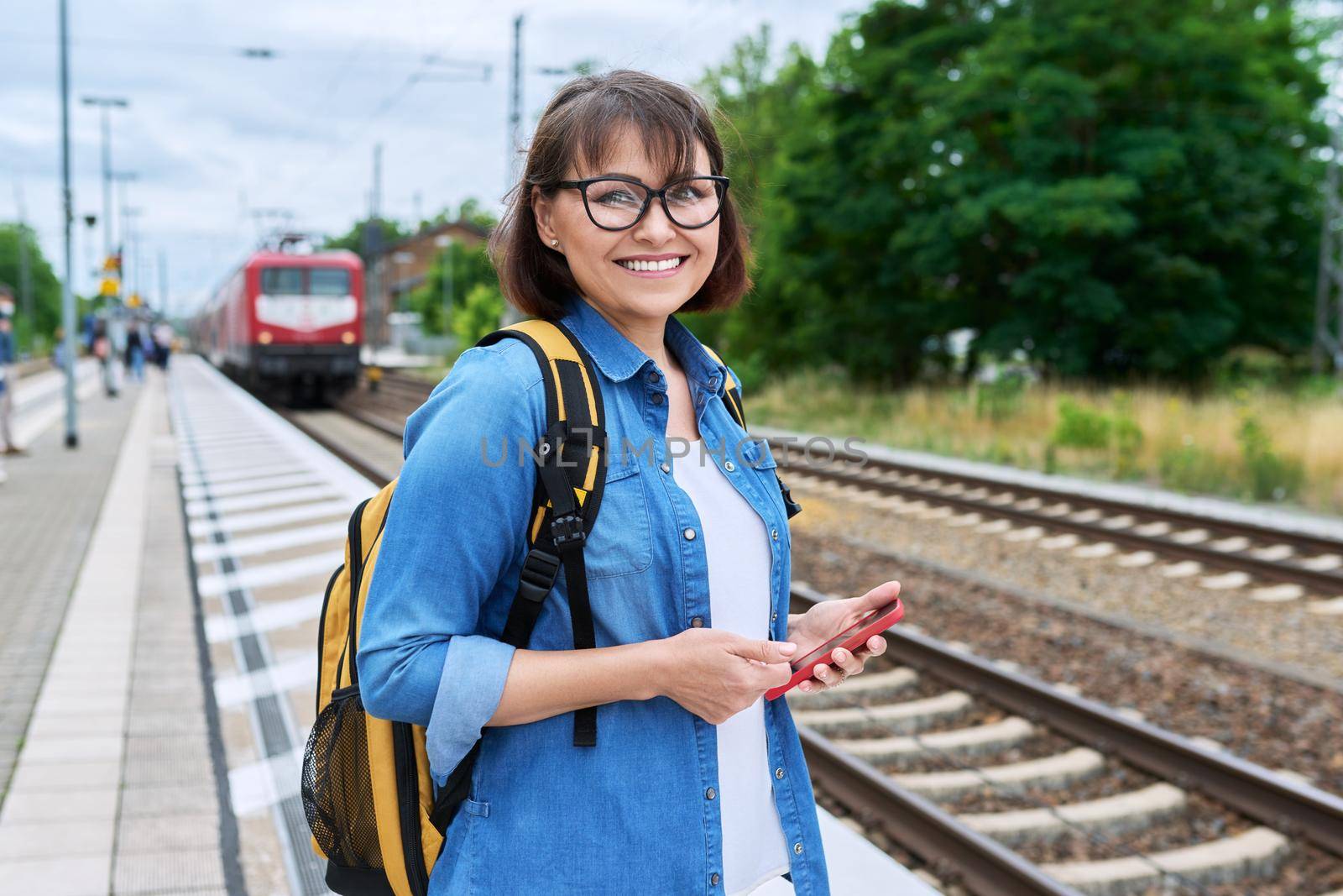 Woman passenger waiting for local train at station outdoor platform, smiling female looking at camera. Rail transport, passenger transportation, journey, tourism, travel, trip, people concept