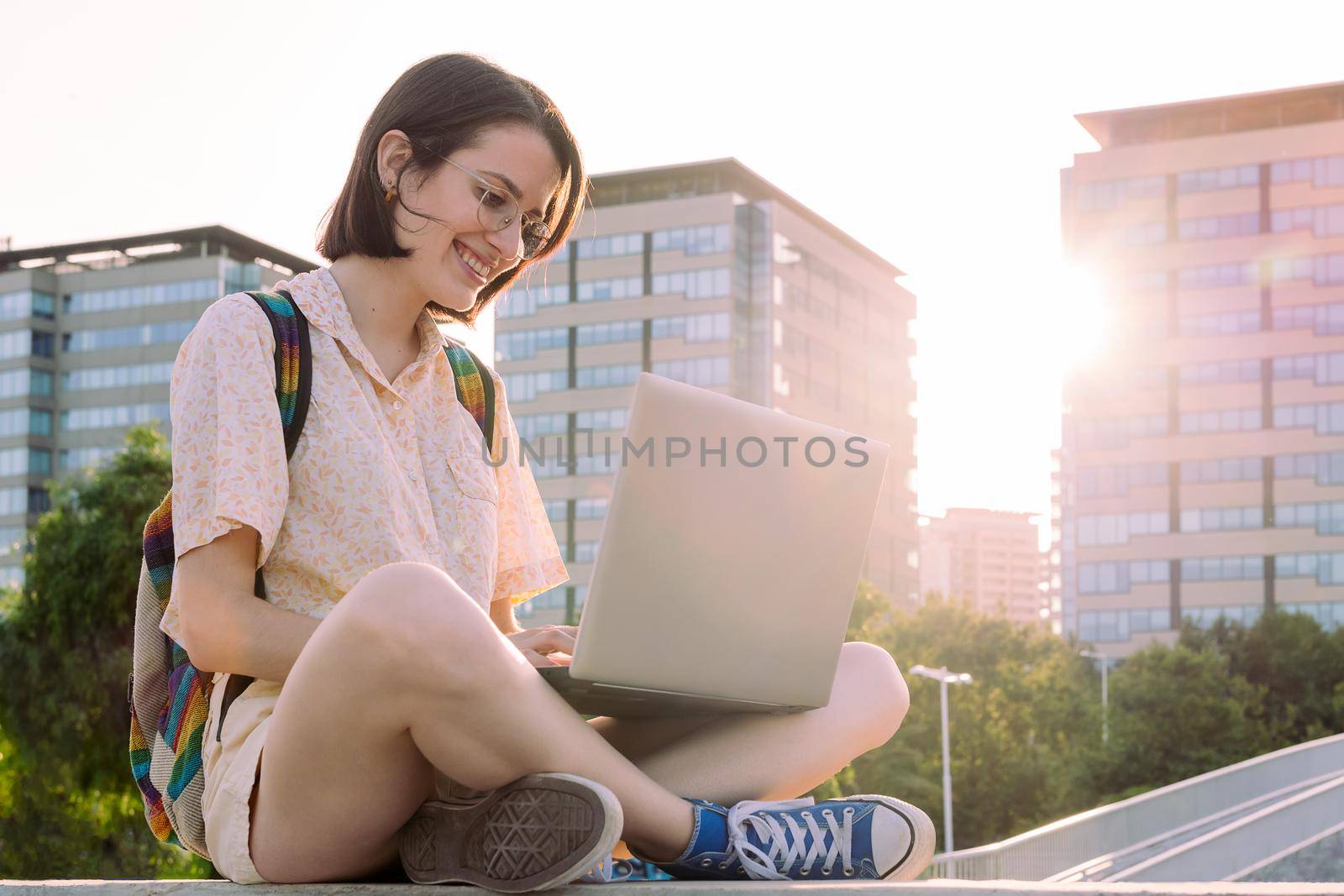 happy young traveler woman with backpack sitting in the city typing on her computer, concept of technology, youth and digital nomad lifestyle, copy space for text
