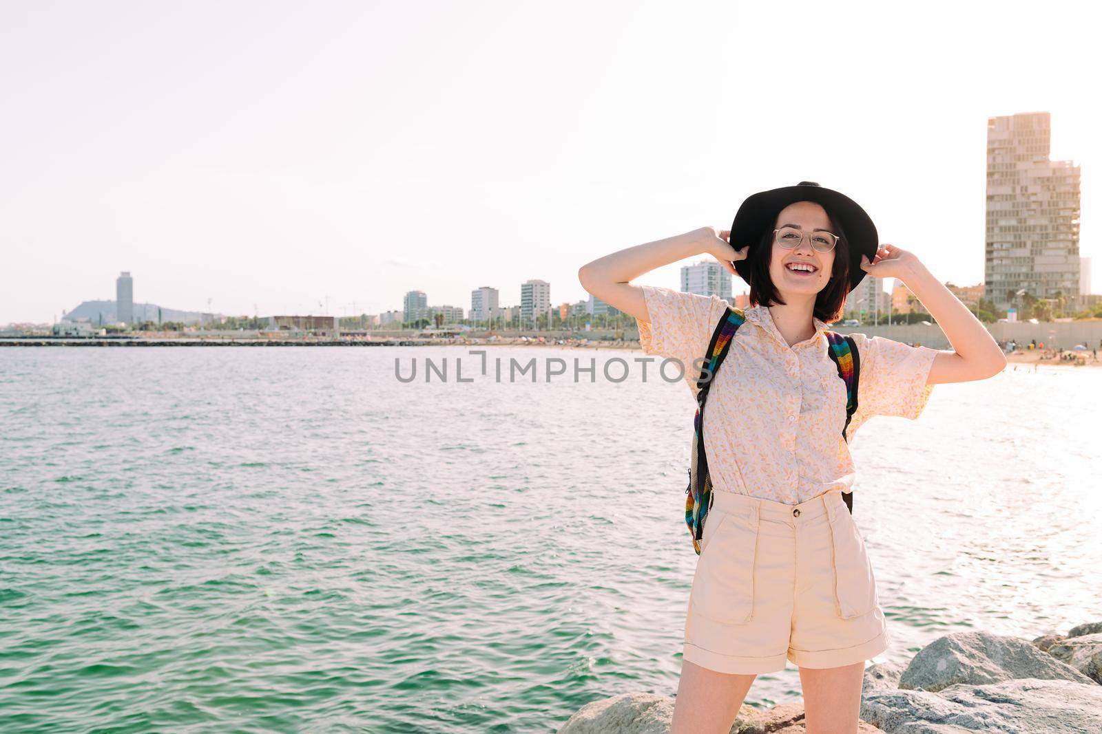 young traveler woman with hat smiles happy near the sea next to the city, travel and tourism concept, copy space for text