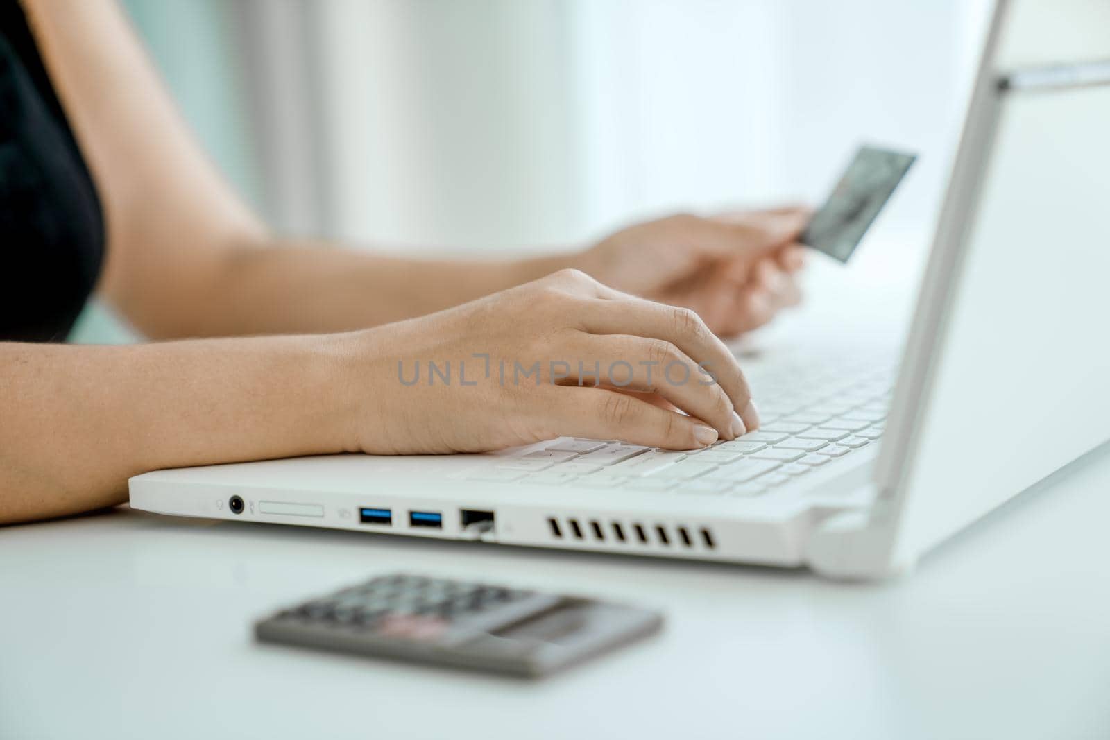 Woman makes online purchases sitting in front of laptop with bank card in her hand. Hands close-up. Concept of online shopping and money transfer.