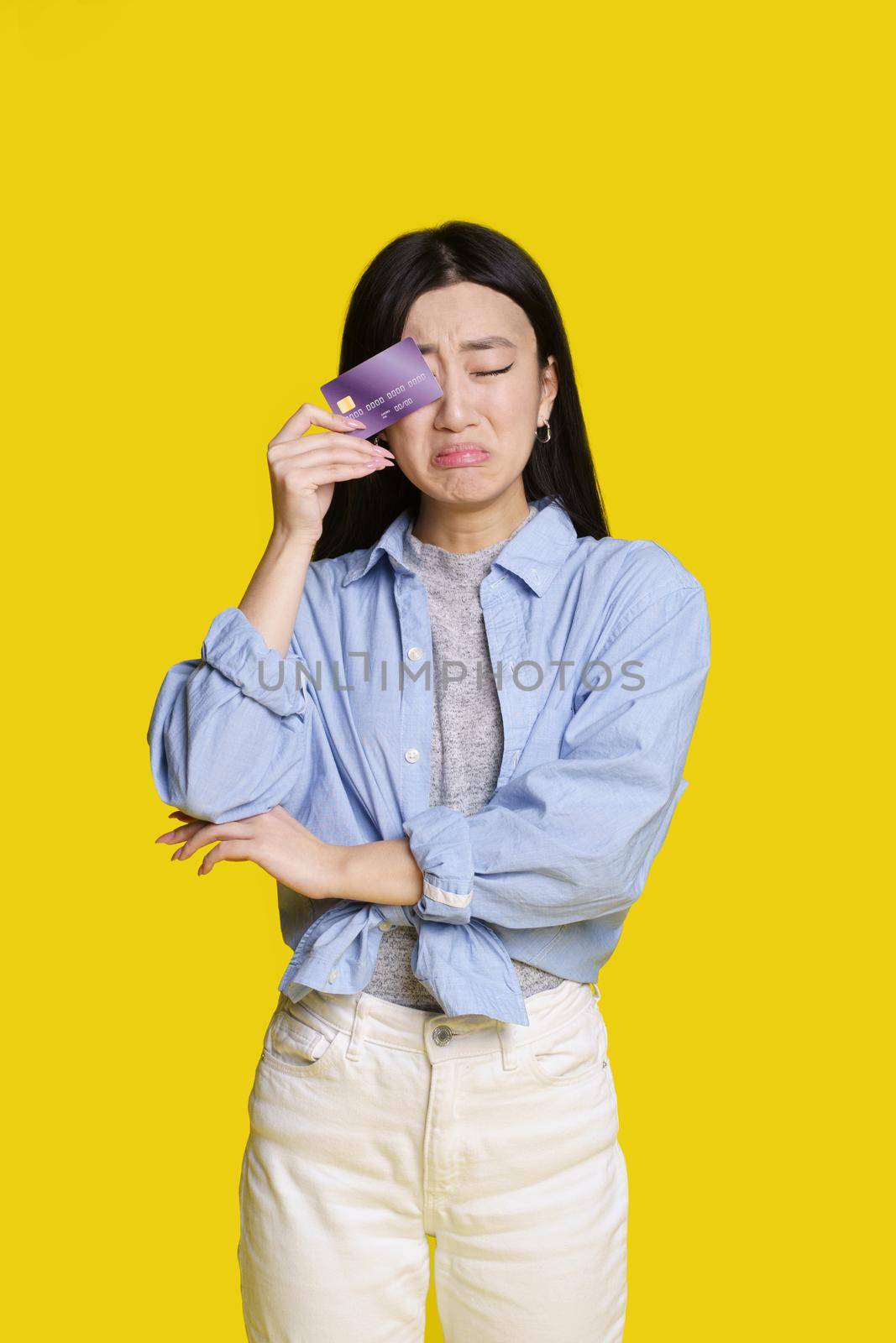 Sad, unhappy asian girl cover her with bank card right eye about to cry that no money. Asian girl in blue shirt with mockup bank card isolated on yellow background. Shopping concept.