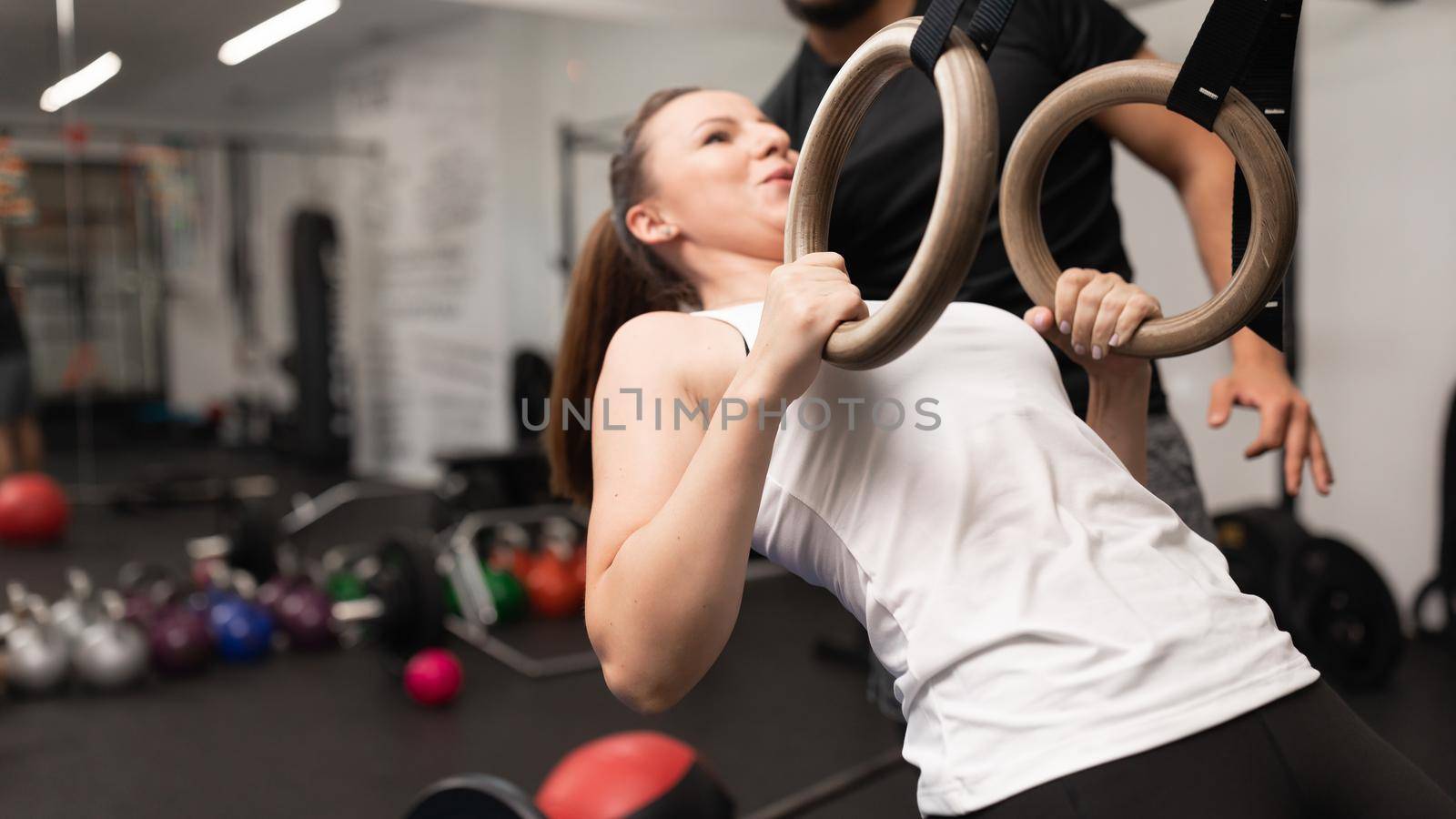 A young woman plays sports and performs single arm ring row and ring hold at the local fitness and training center with her personal trainer