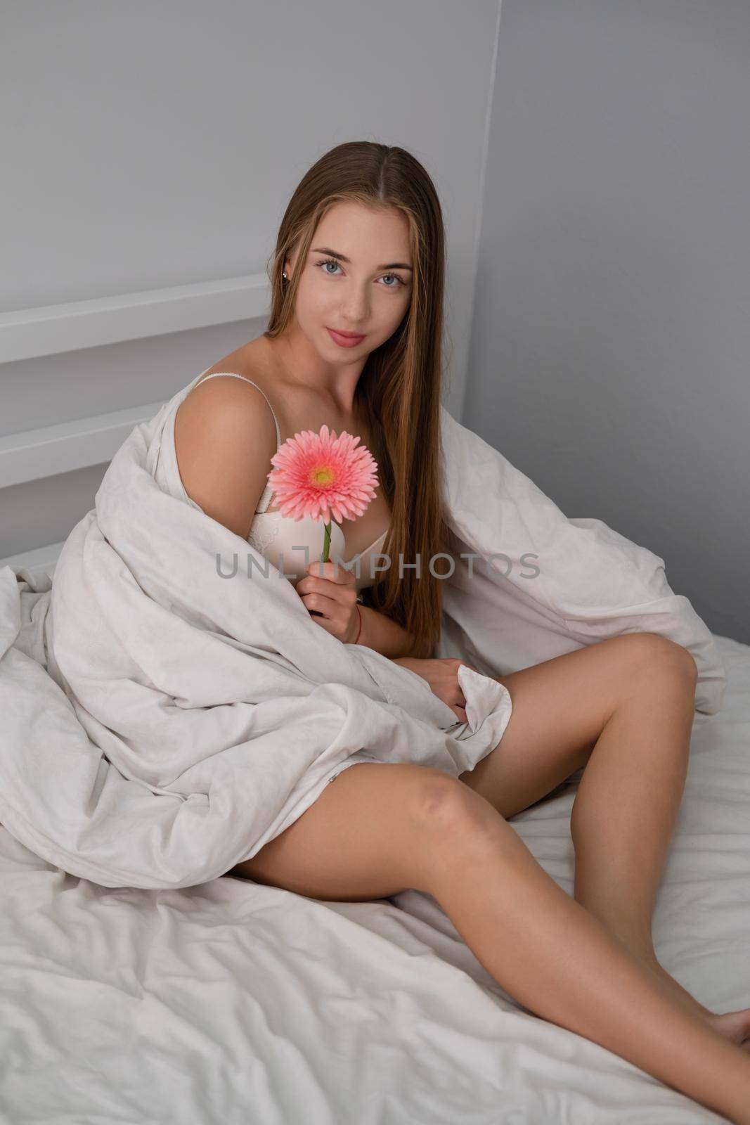 charming young woman lies in underwear on comfortable bed. holding red pink flower. tender portrait of a girl in a hotel room or home. Lovely female enjoy good relaxation. Morning time.
