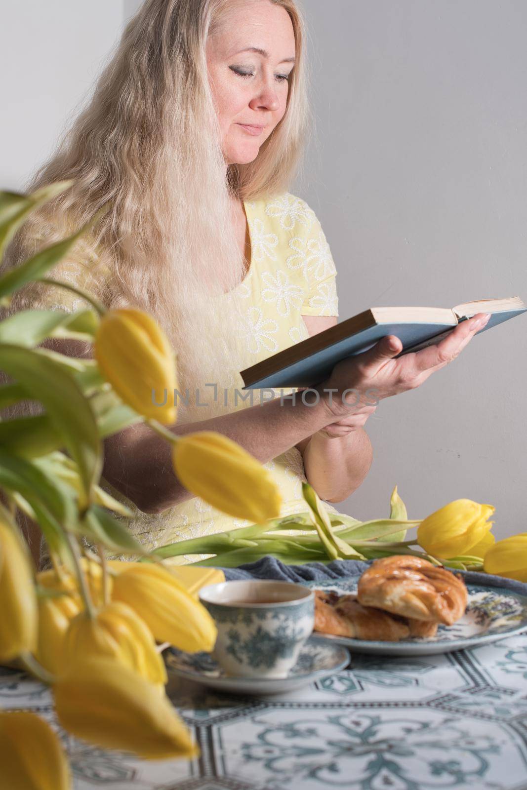 a middle-aged woman reads a book at a table with a dessert and yellow tulips, by KaterinaDalemans