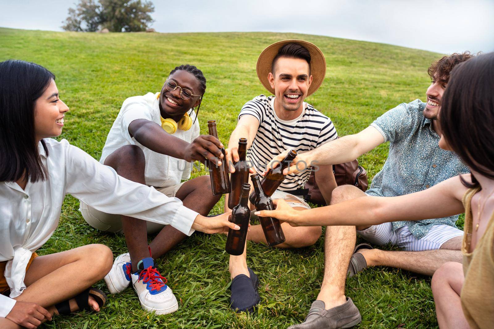 Multiracial happy and smiling friends having fun together in a park toasting with beers. Friendship concept.