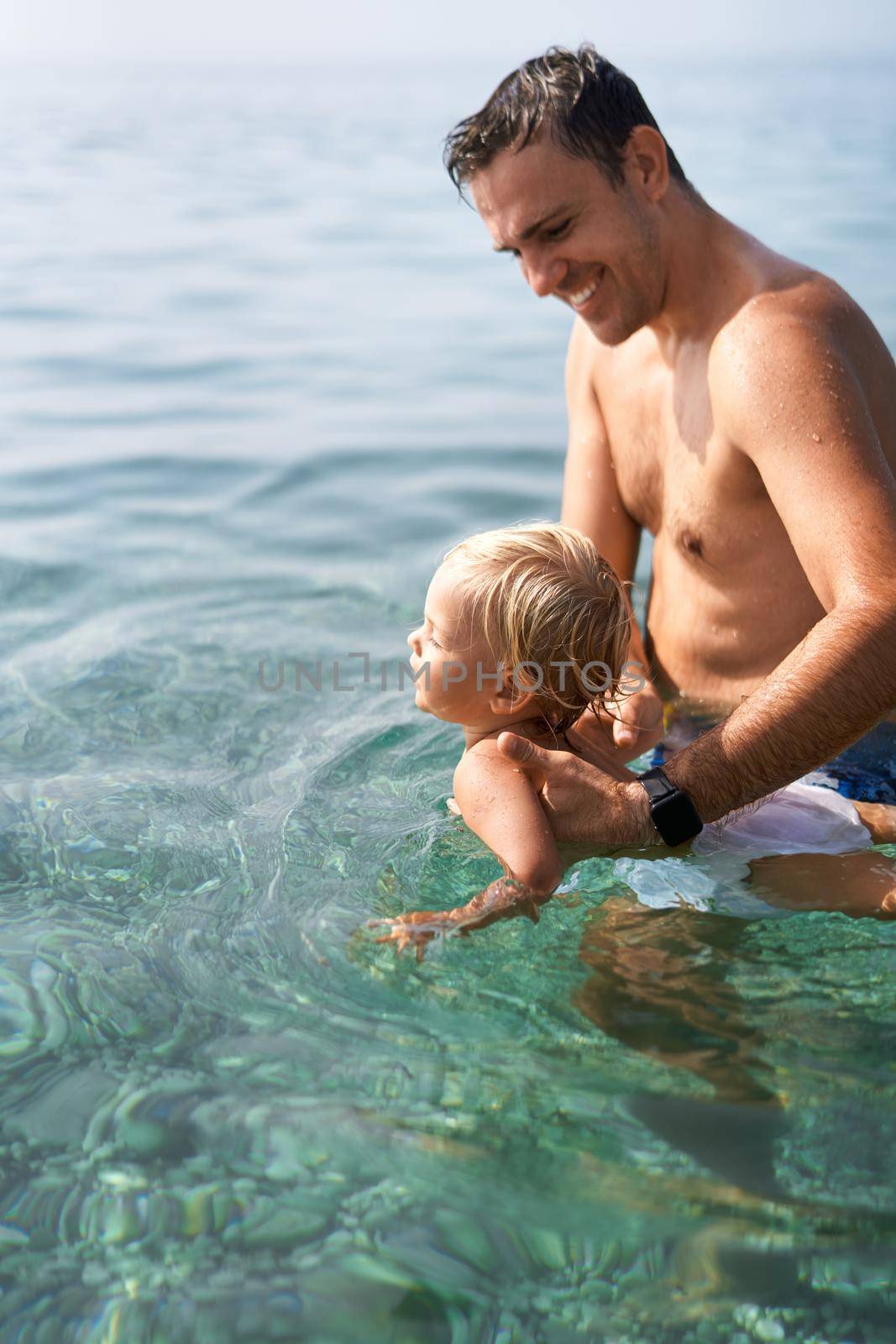 Smiling dad teaches little daughter to swim holding her hands under armpits by Nadtochiy