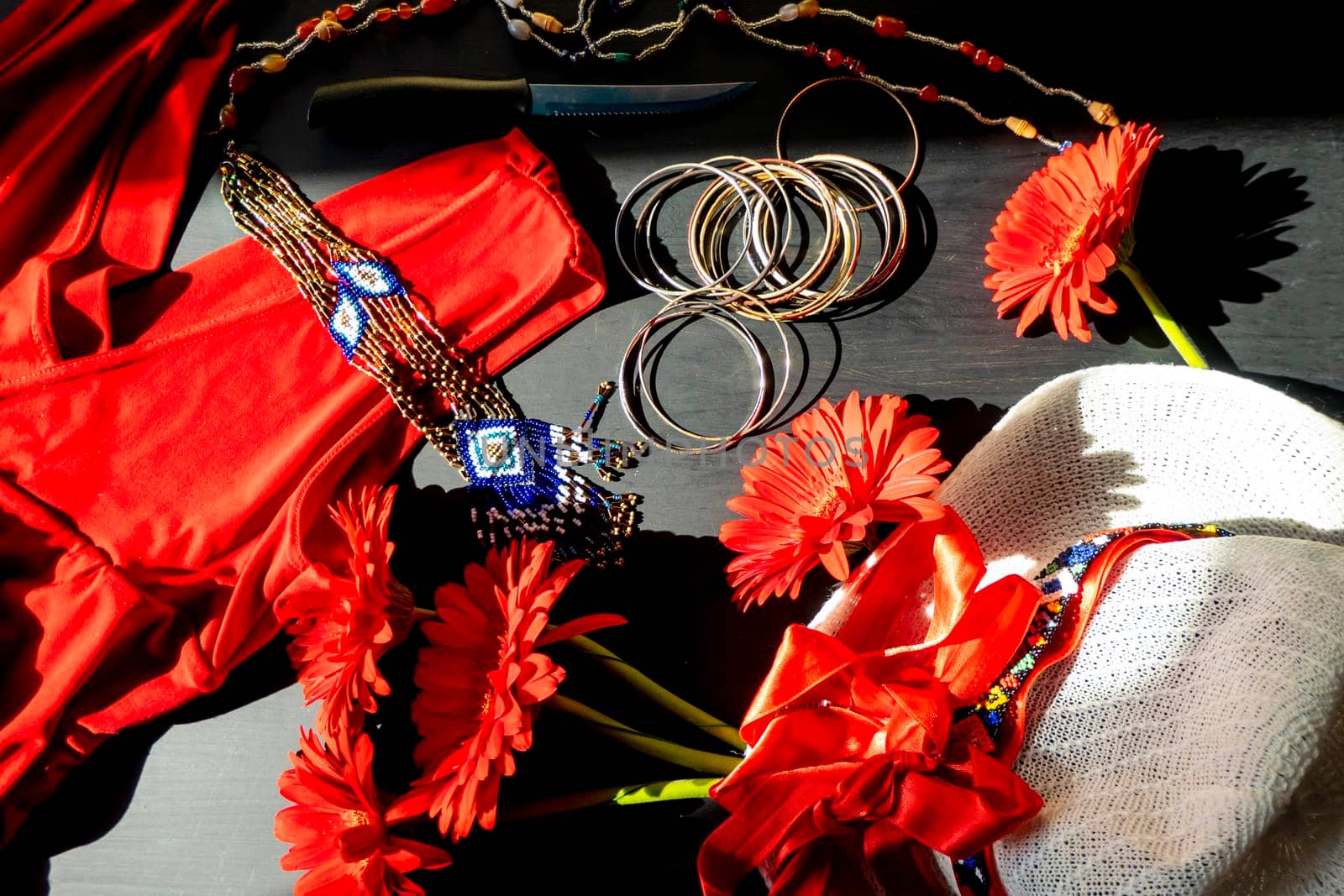 flat lay of a fashionable look from red and white accessories on a black background. Scarlet dress and white hat with a ribbon, jewelry made of beads and small stones, light jingling bracelets