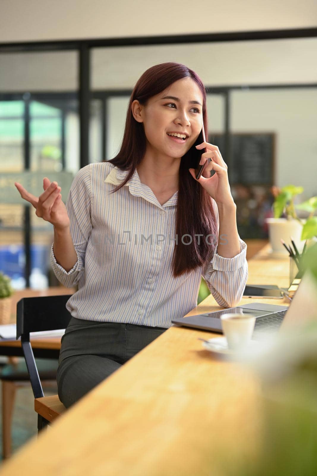 Shot of young female entrepreneur having pleasant phone conversation with her business partner, sitting in coffee shop.