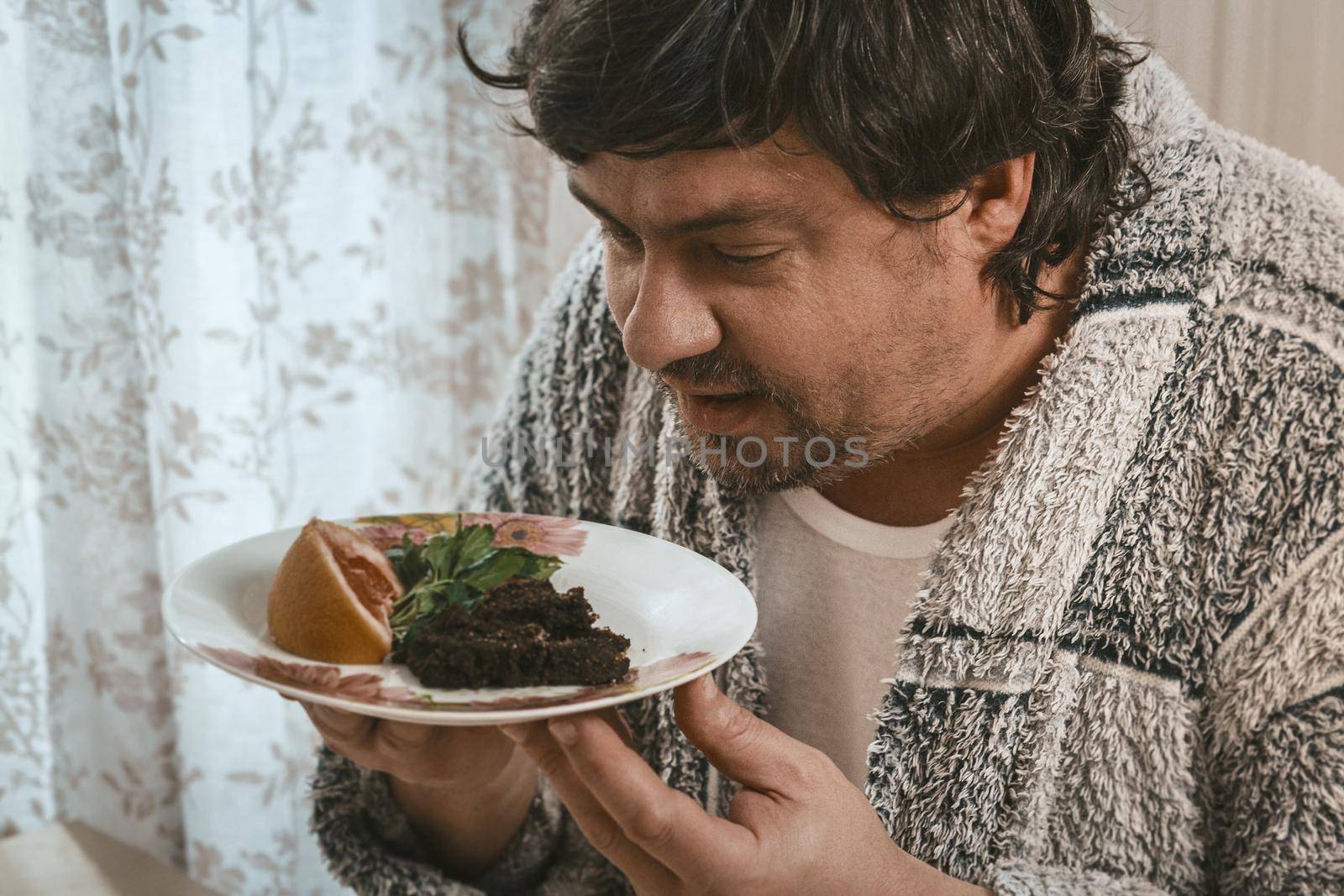 Body Positive Man Looks At A Plate With Wholesome Food, Sleepy Caucasian Man In Home Clothes Holds A Plate With Salad And Grapefruit In His Hands