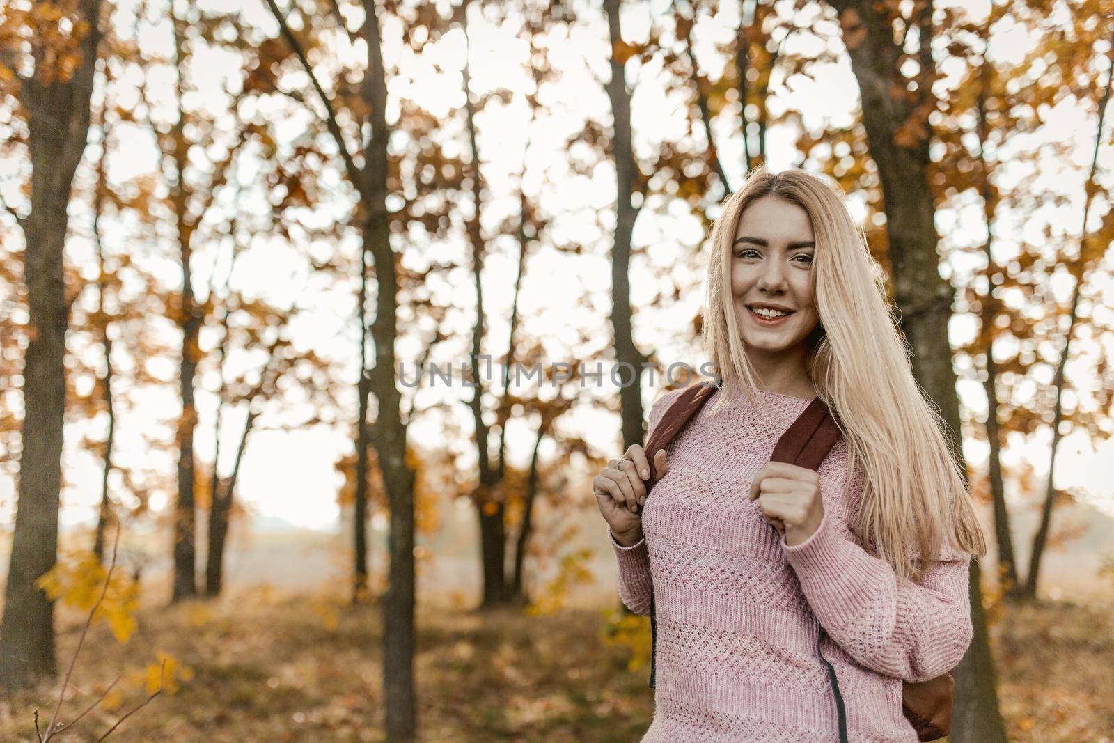 Young Female Traveler Posing On Background Of Autumn Forest Outdoors, Charming Blonde With Backpack Stopped Looking At Camera, Autumn Trees With Yellow Leaves On Sunny Blurry Background